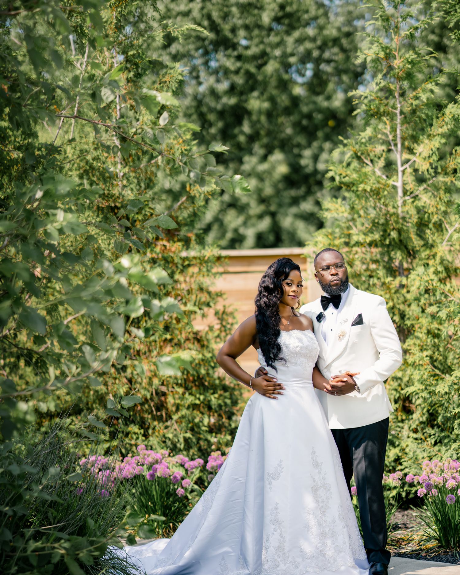 Black bride and groom in white wedding attire pose elegantly among lush garden greenery.