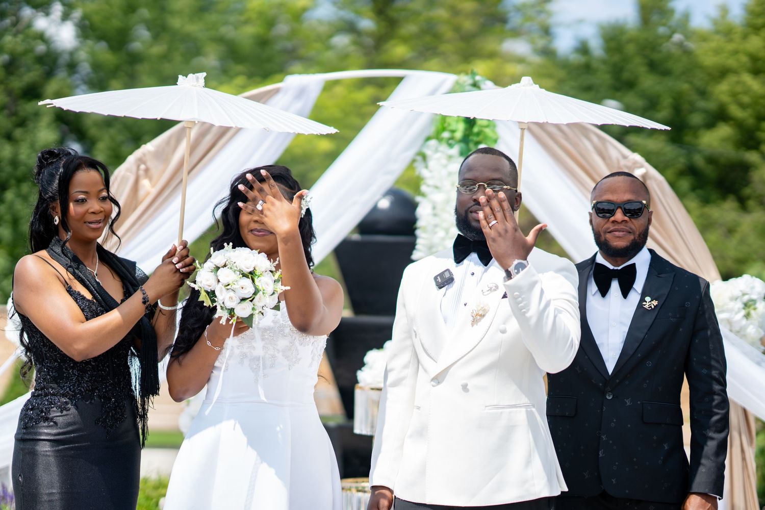 Emotional groom wiping tears at outdoor wedding ceremony with bride in white gown and wedding party.