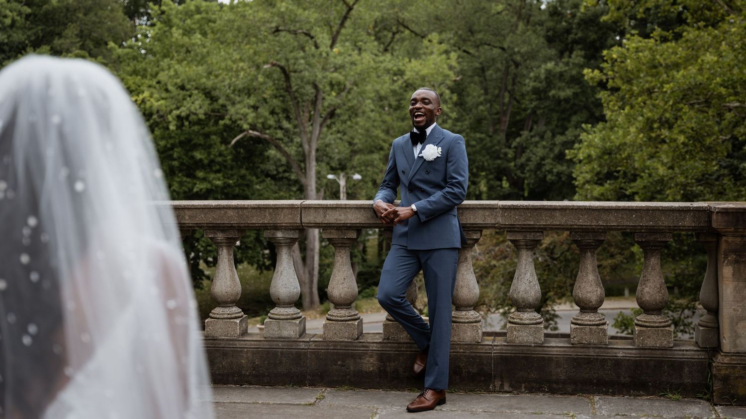 Groom in blue suit smiles seeing bride in white veil during wedding first look outdoor ceremony.