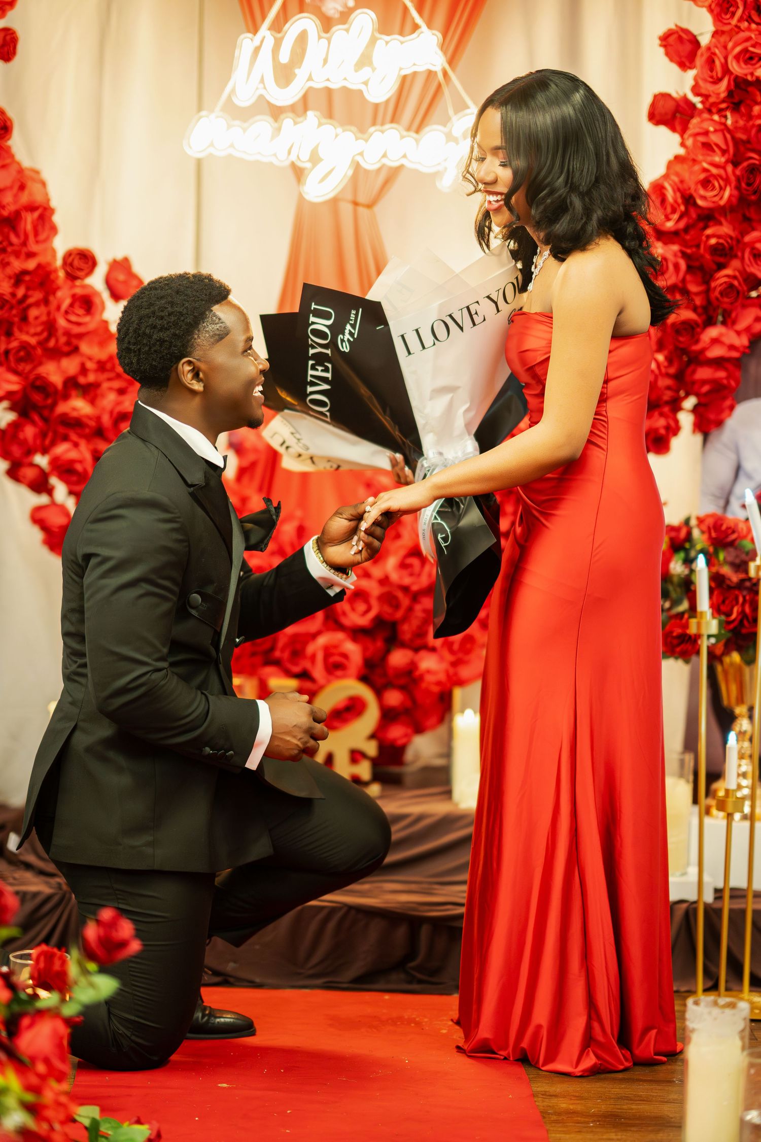 A man kneeling proposes to a woman in a red gown holding flowers with a Will You Marry Me neon sign backdrop.