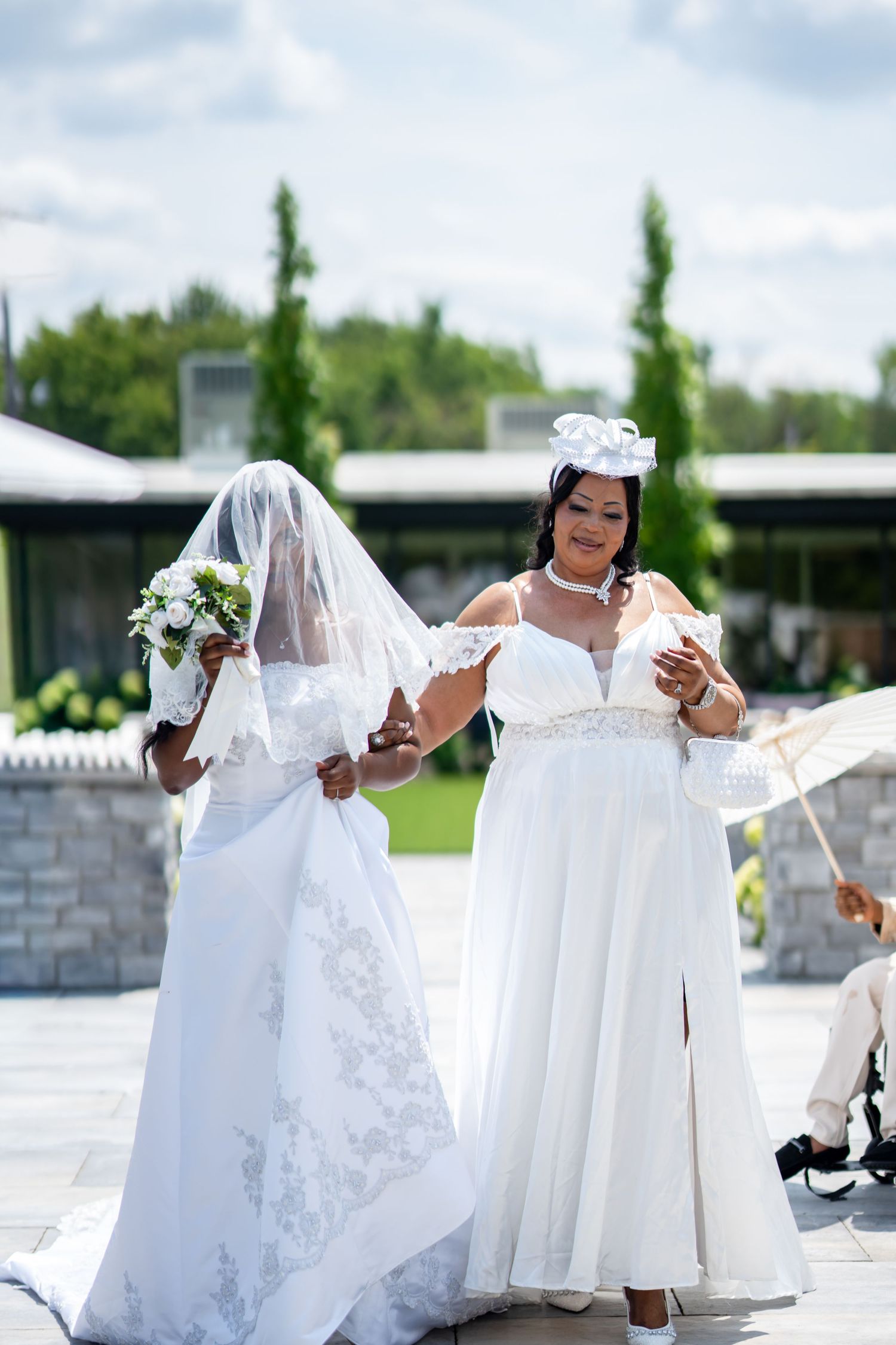 Bride in lace wedding gown with veil walking alongside smiling bridesmaid in white dress outdoors.