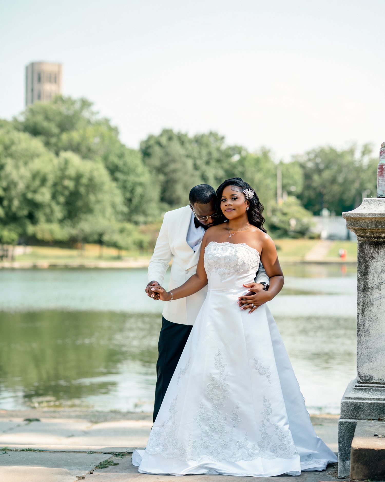 Elegant bride and groom posing by a serene waterfront, bride in strapless lace gown, groom in white tuxedo jacket.