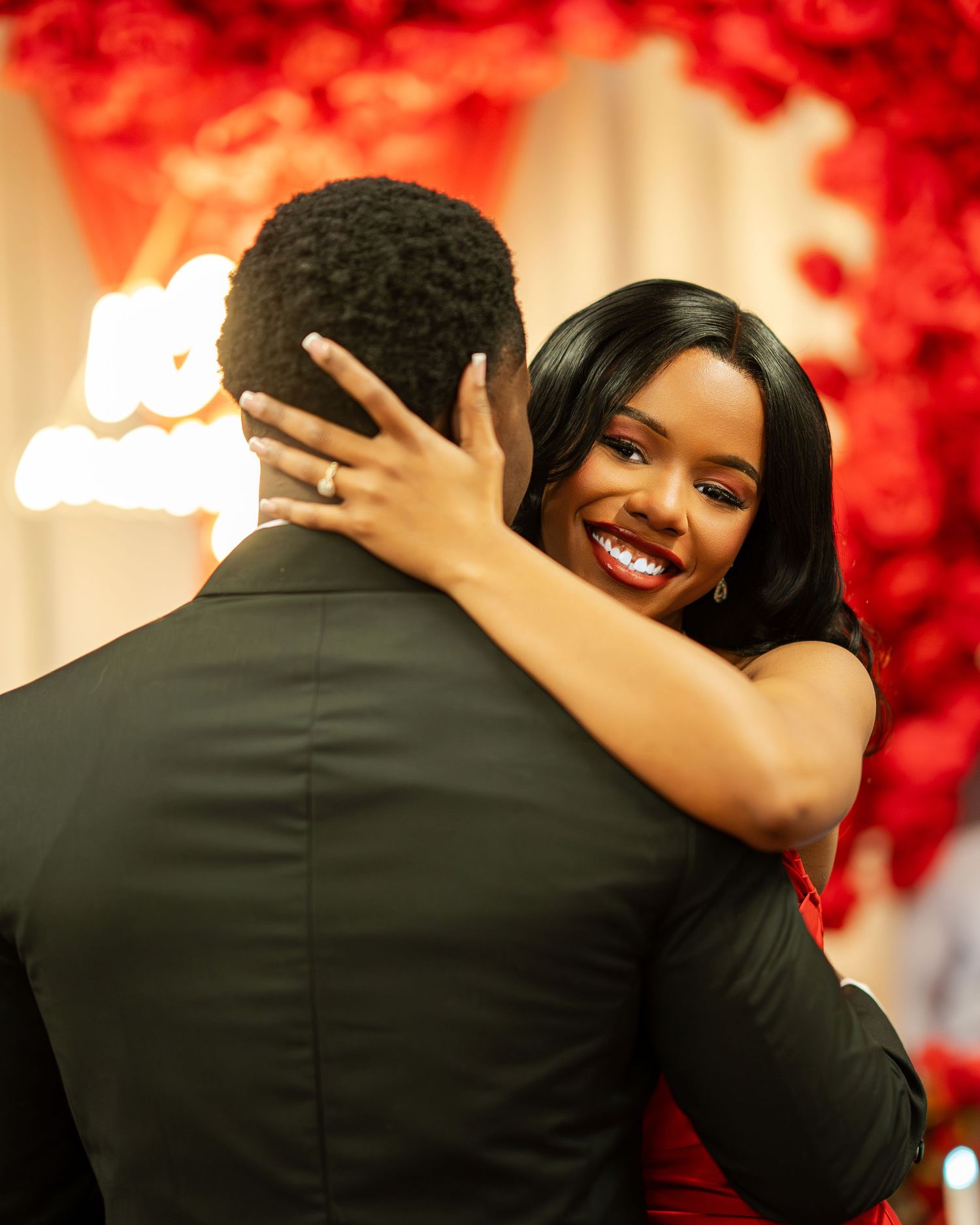 A smiling woman in a red dress embraces a man in a black suit during a romantic red rose-filled proposal event.