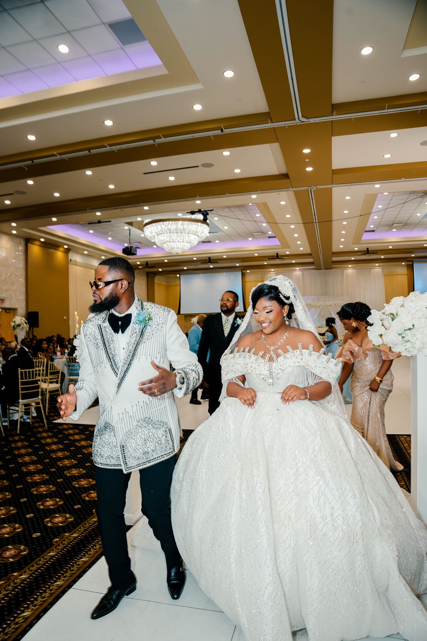 Bride in white ballgown and groom in silver tuxedo jacket make grand entrance at wedding reception.