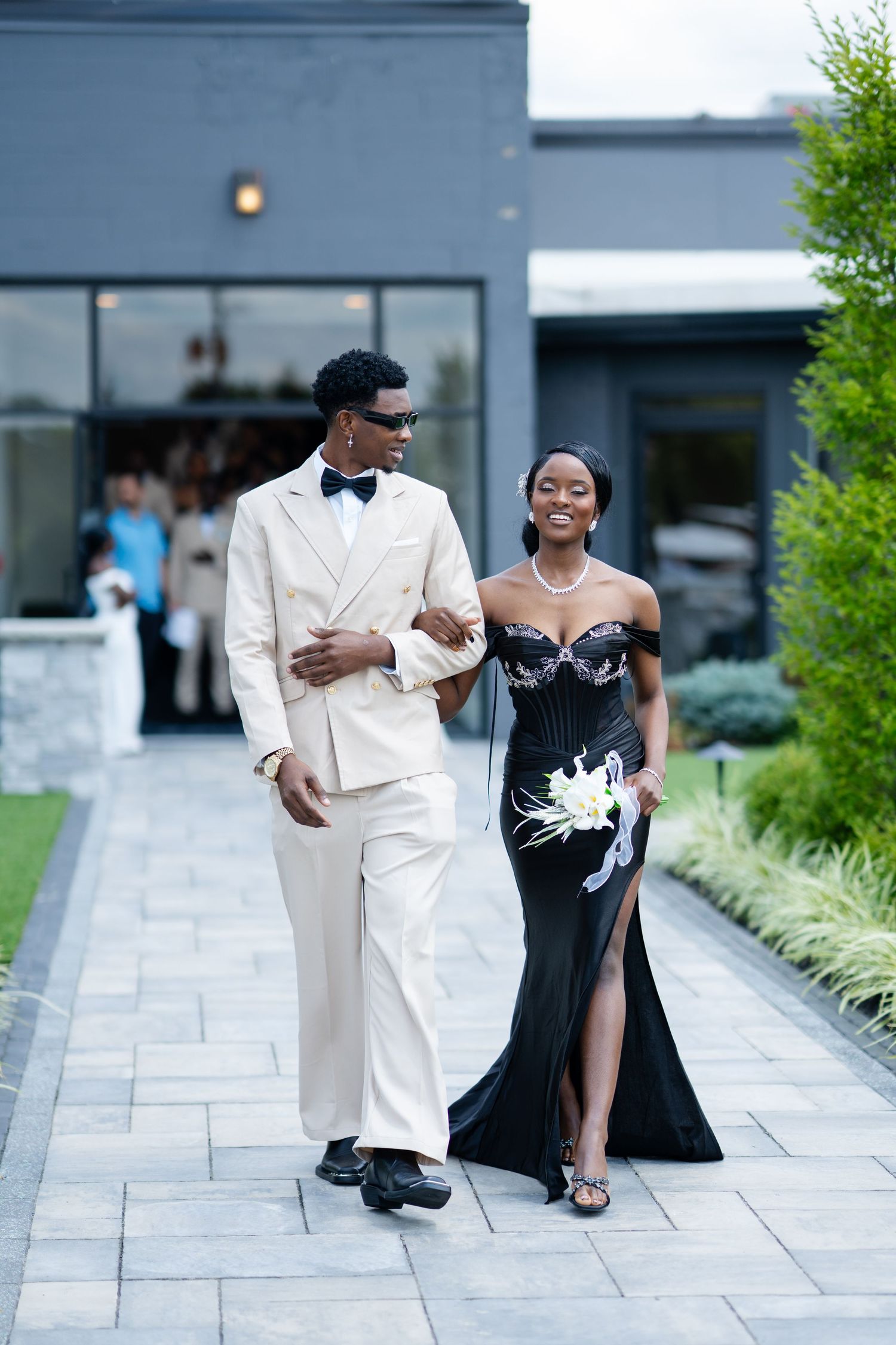 Elegant couple walking outdoors, man in cream suit and woman in black off-shoulder gown with floral detail.