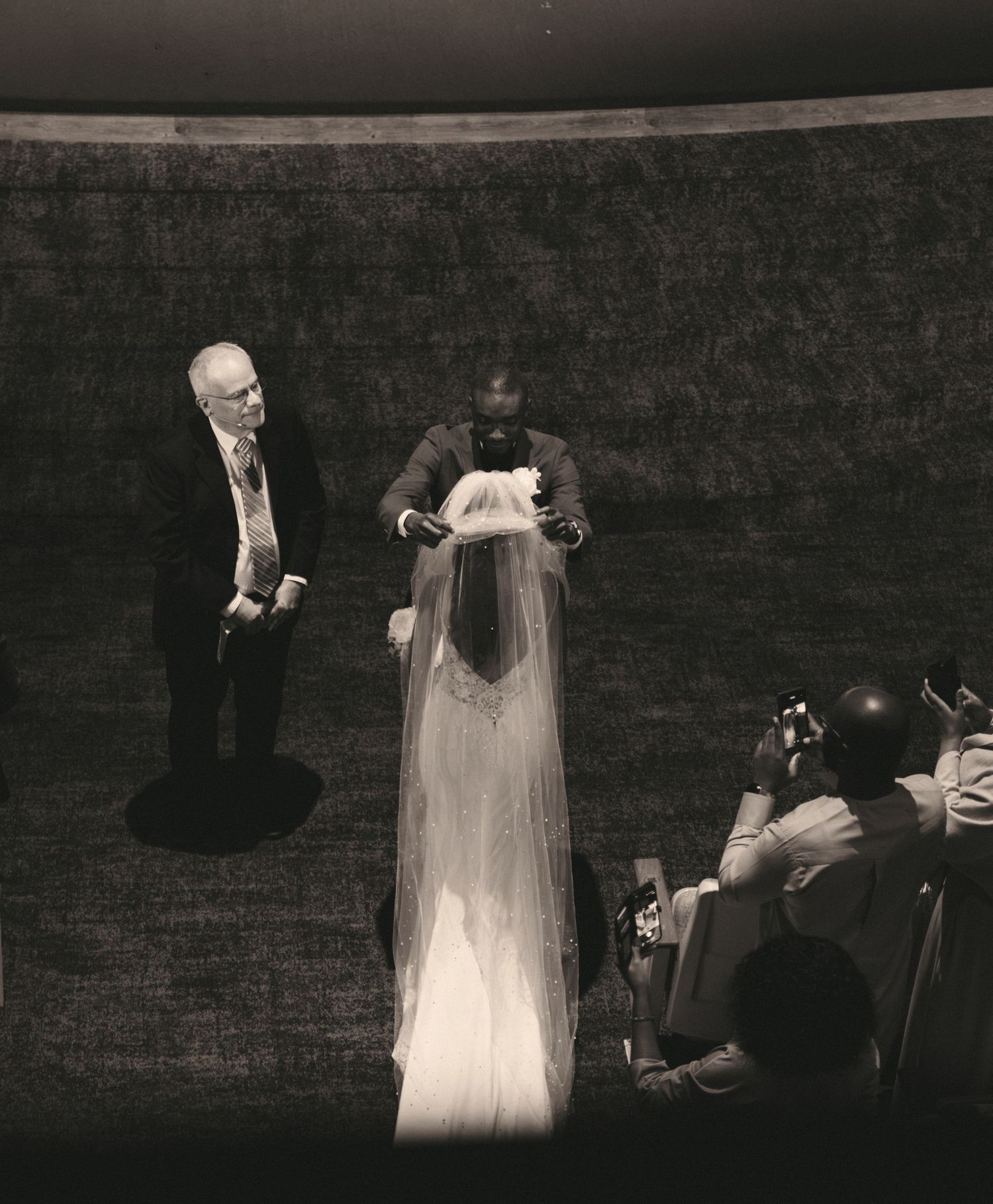 Black and white photo of groom lifting bride veil during intimate indoor wedding ceremony with officiant.