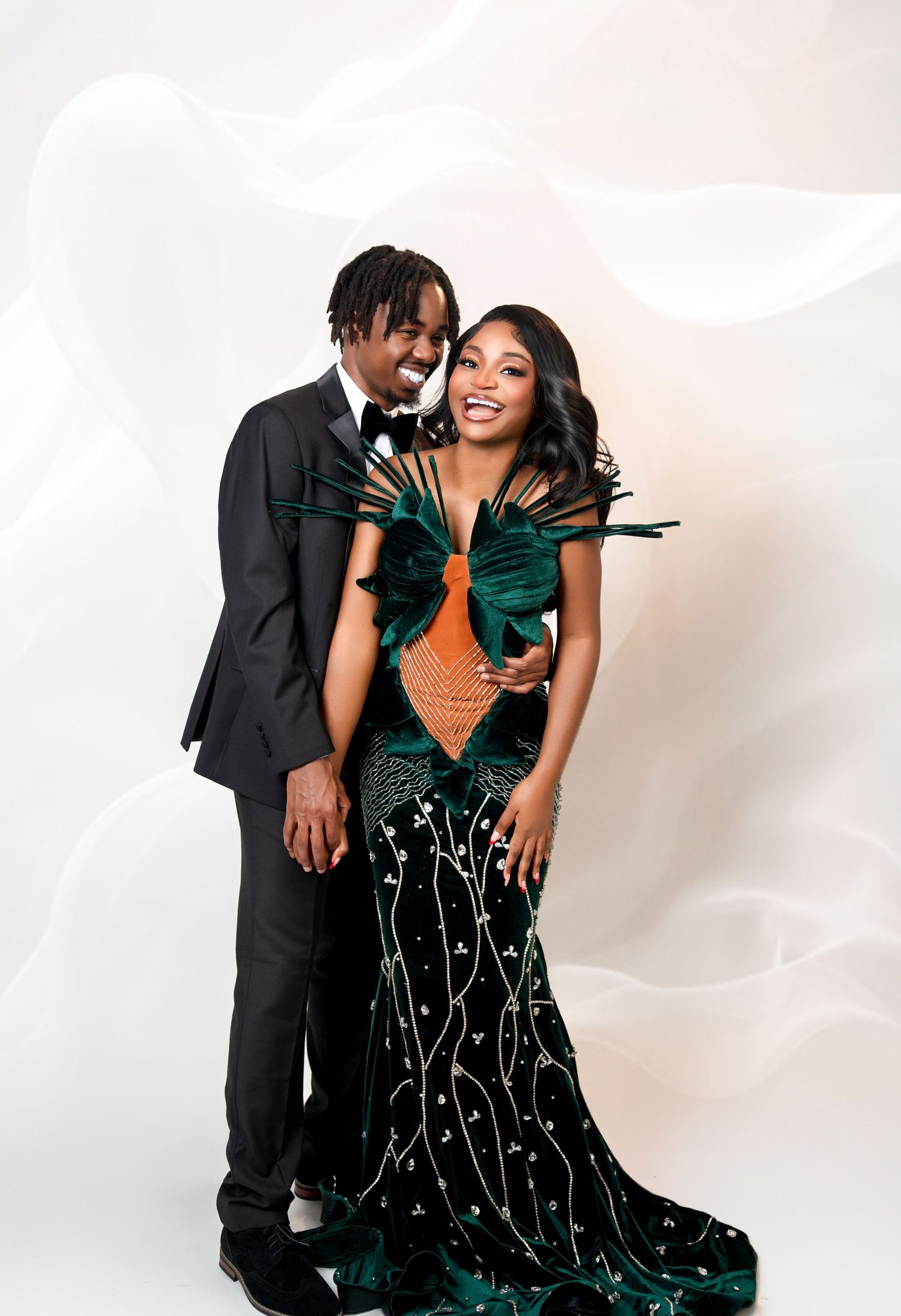 A Black couple poses elegantly, wearing a stunning green feathered gown and black tuxedo in a studio.