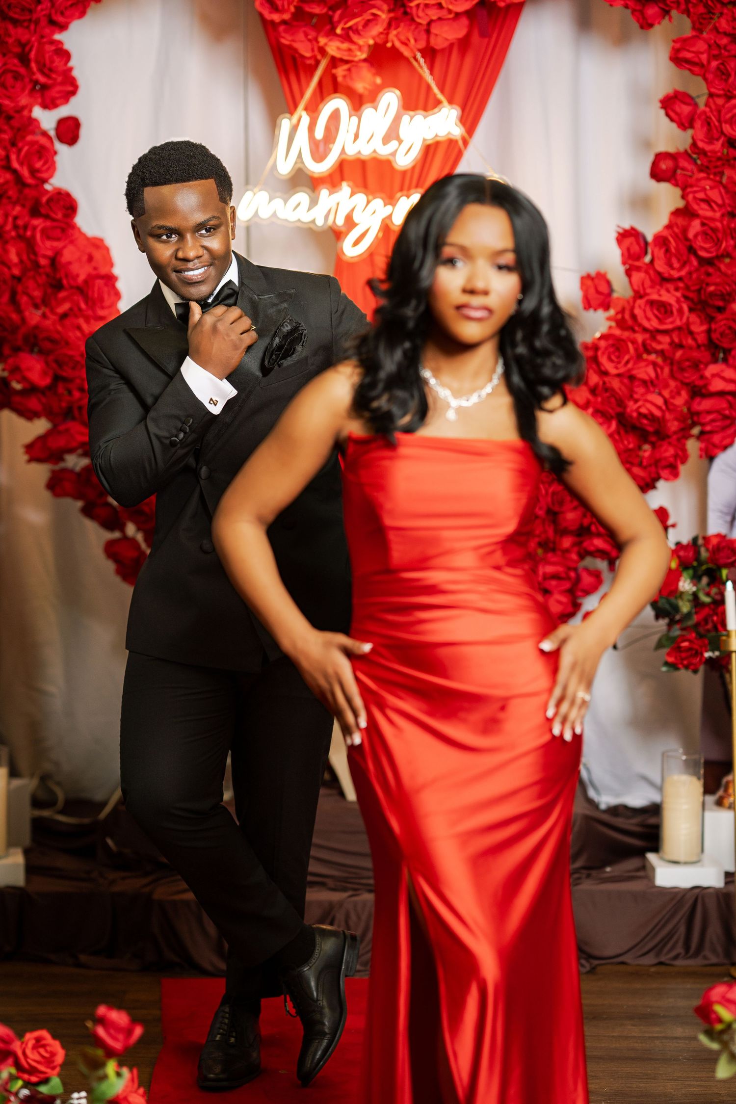 A stylish couple poses in front of a Will You Marry Me neon sign surrounded by lush red rose decorations.