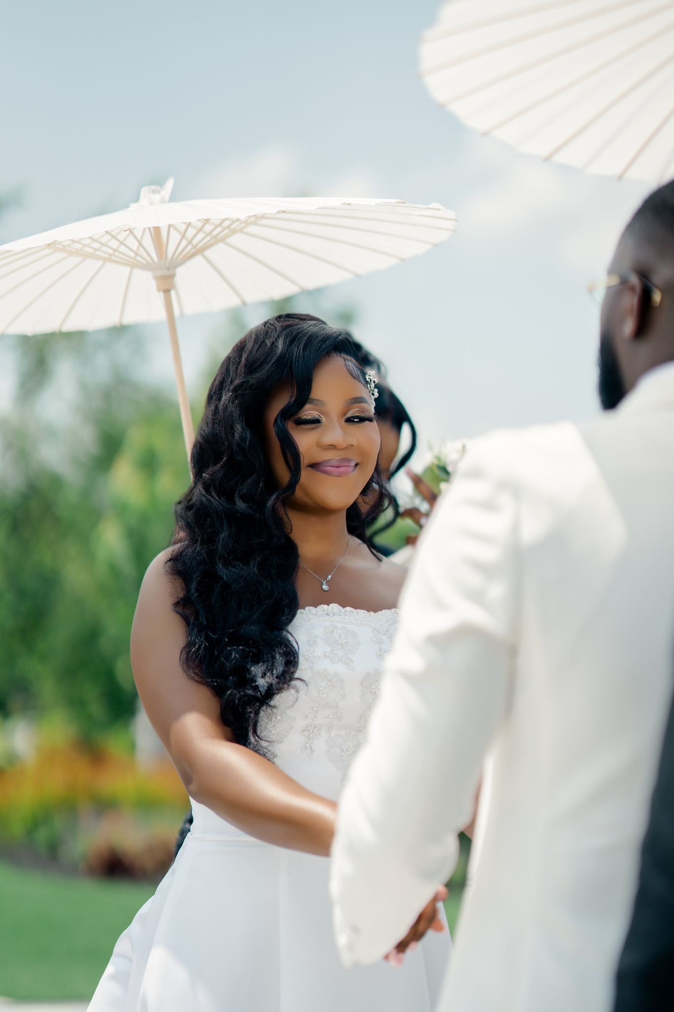 A smiling bride in a strapless white gown holds a parasol while facing her groom during an outdoor wedding ceremony.