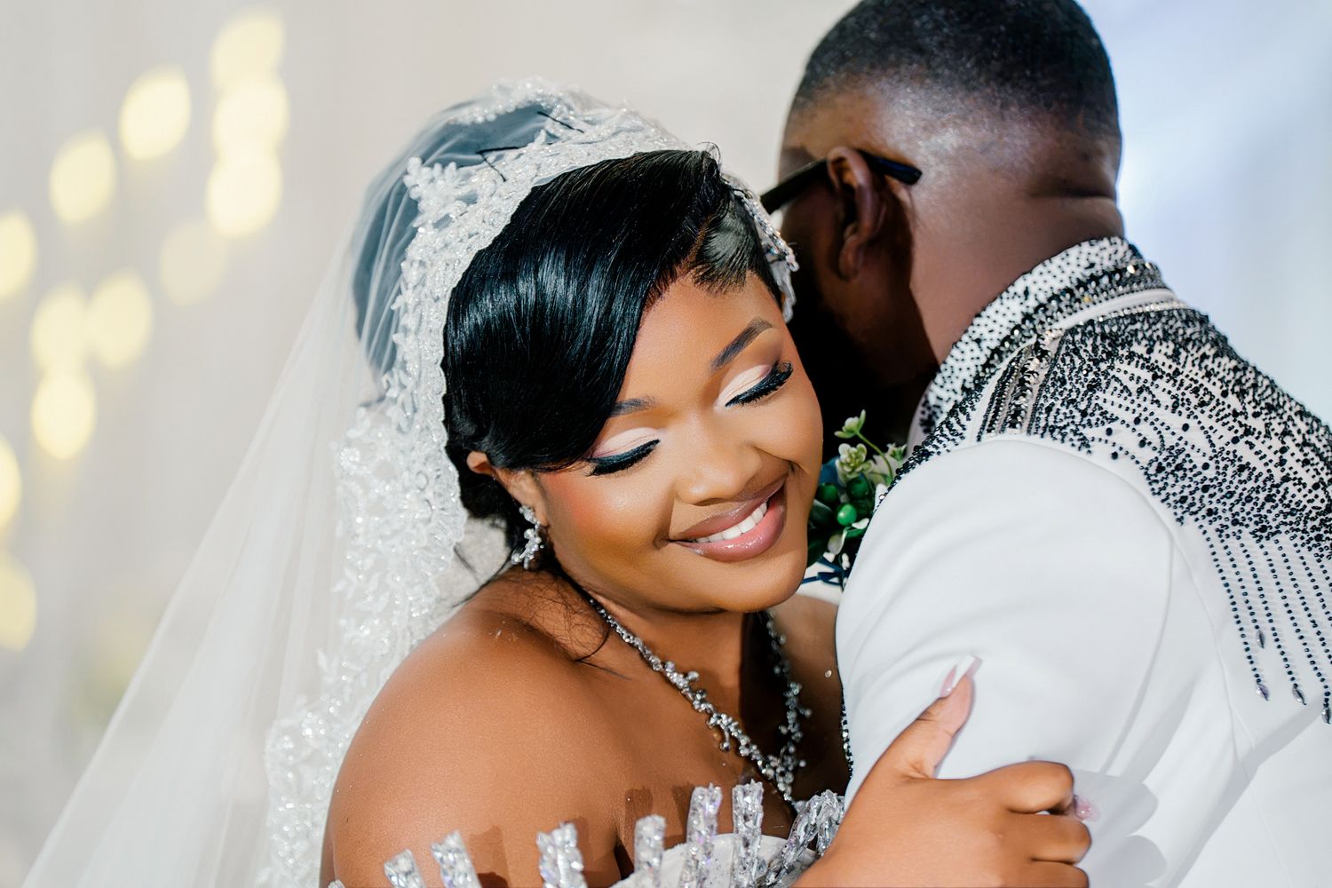 Radiant bride in beaded white gown smiles as groom whispers in her ear at elegant wedding.