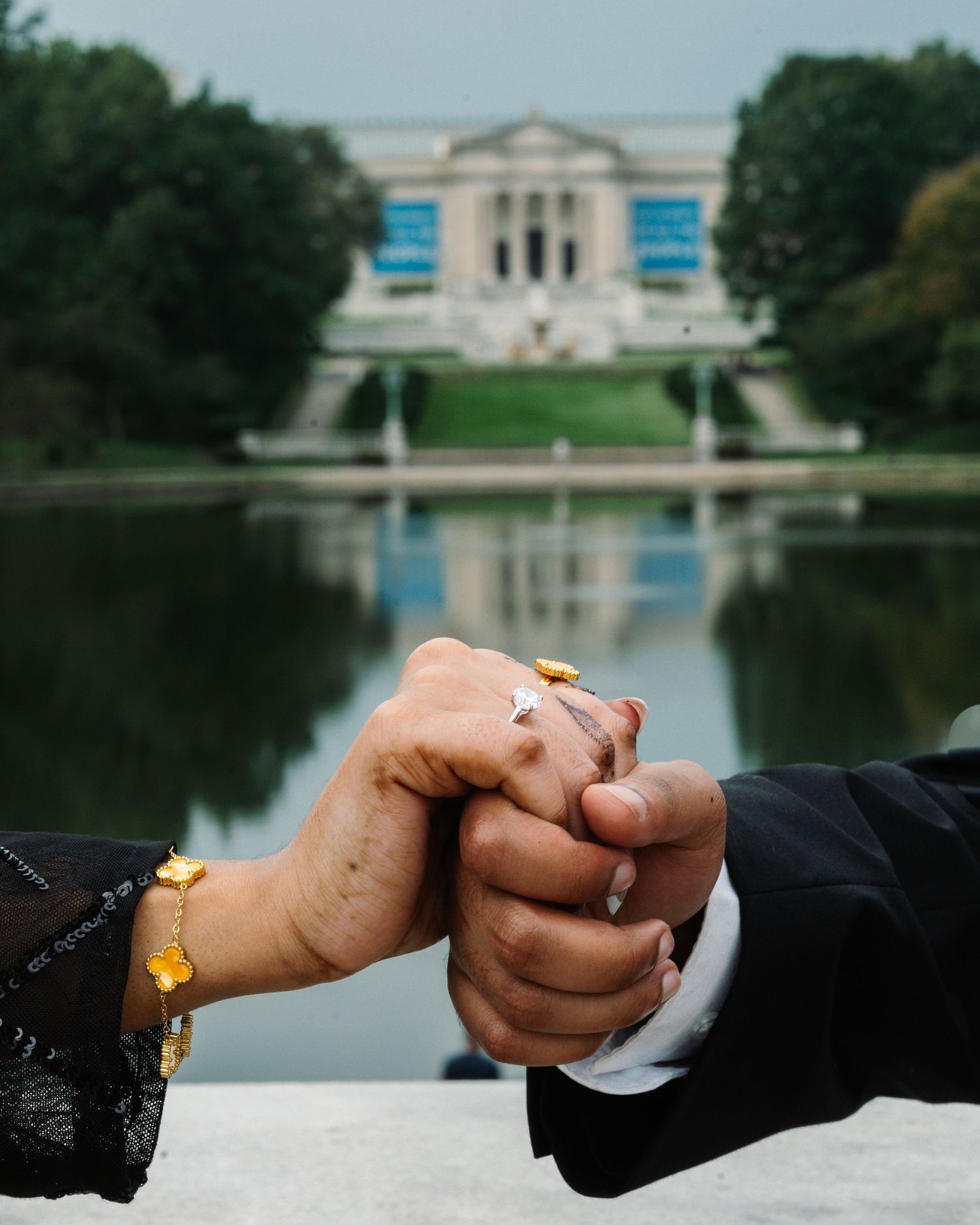 Close-up of two hands clasped together with a ring, in front of a reflecting pool.