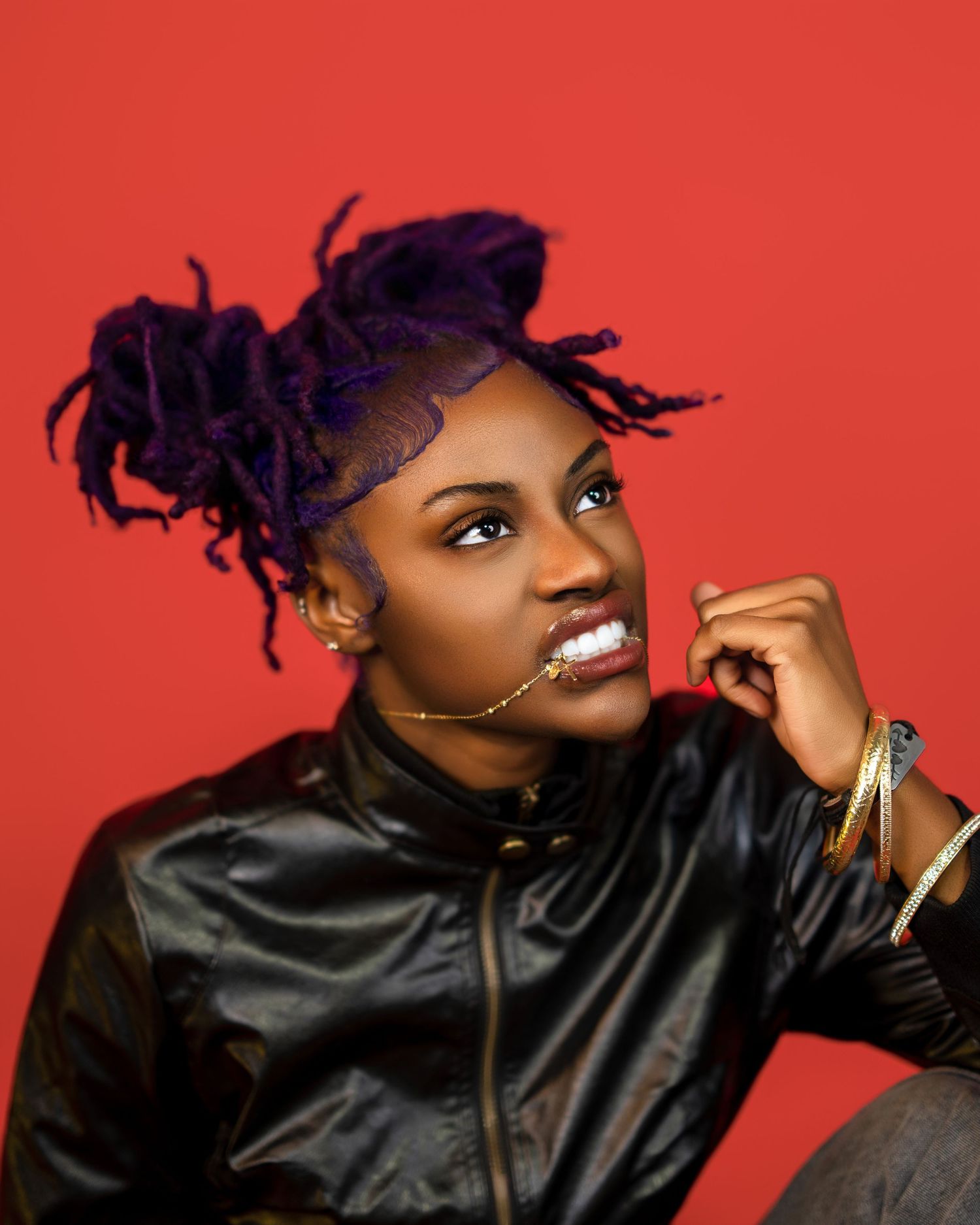 Woman with purple locs poses confidently in a black leather jacket against a bold red background.