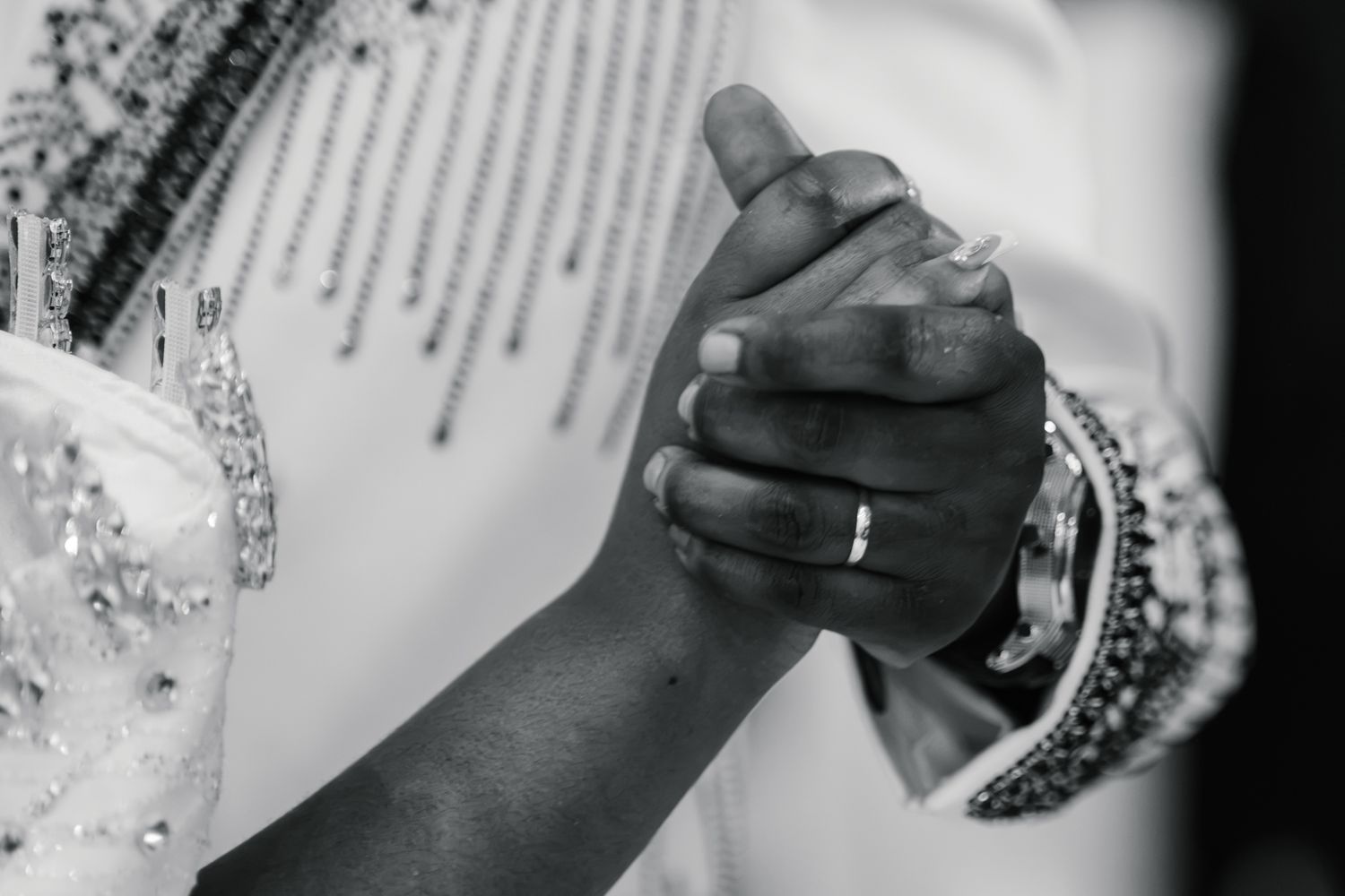 Black and white close-up of bride and groom holding hands showing wedding ring and bridal bracelet.