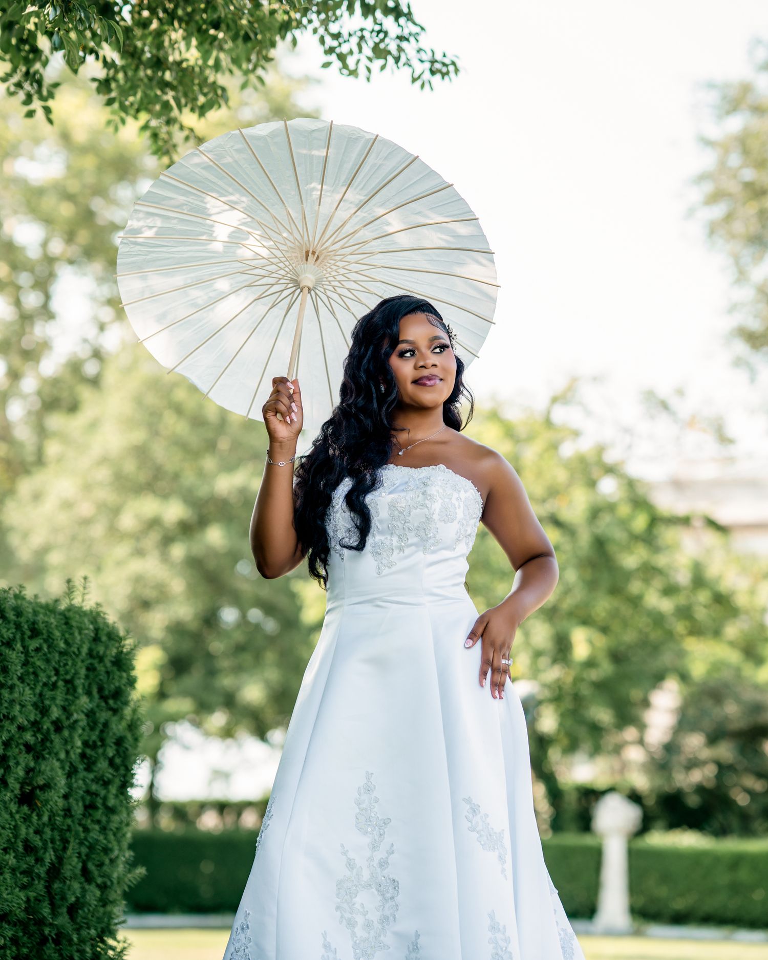 A beautiful bride in a strapless white wedding gown holds a white parasol while posing in a garden setting.