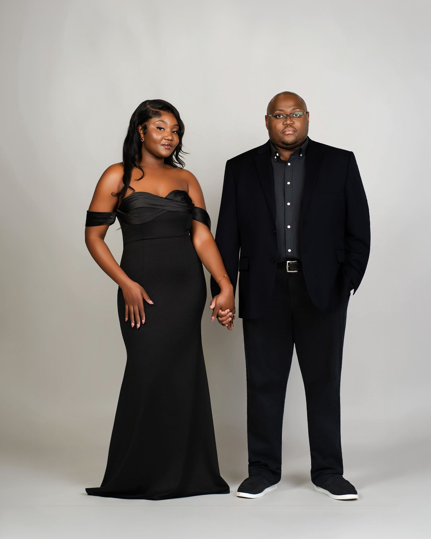 Couple posing together in matching black formal attire holding hands in a studio setting.