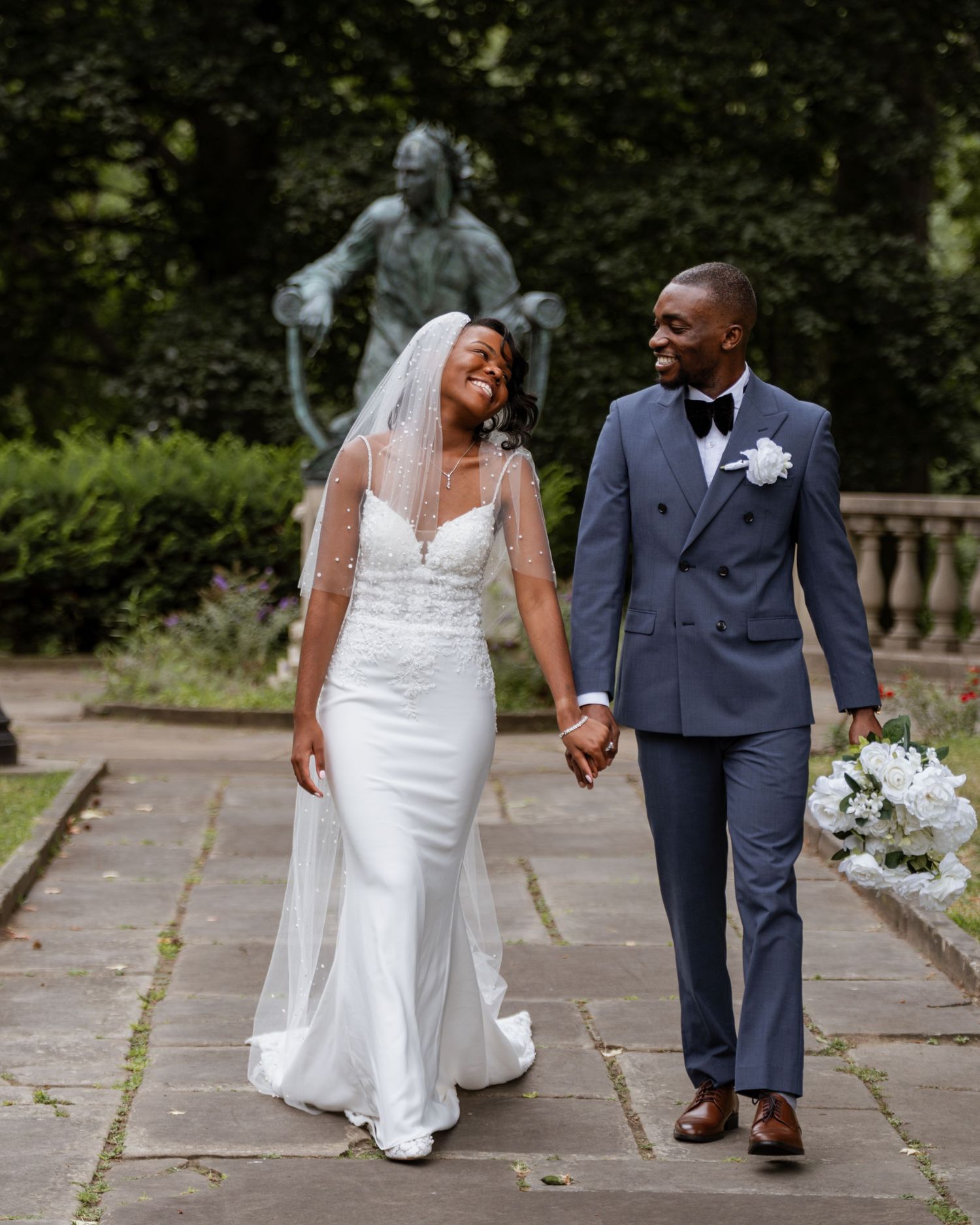 Smiling bride and groom walking hand in hand past a statue in a park.