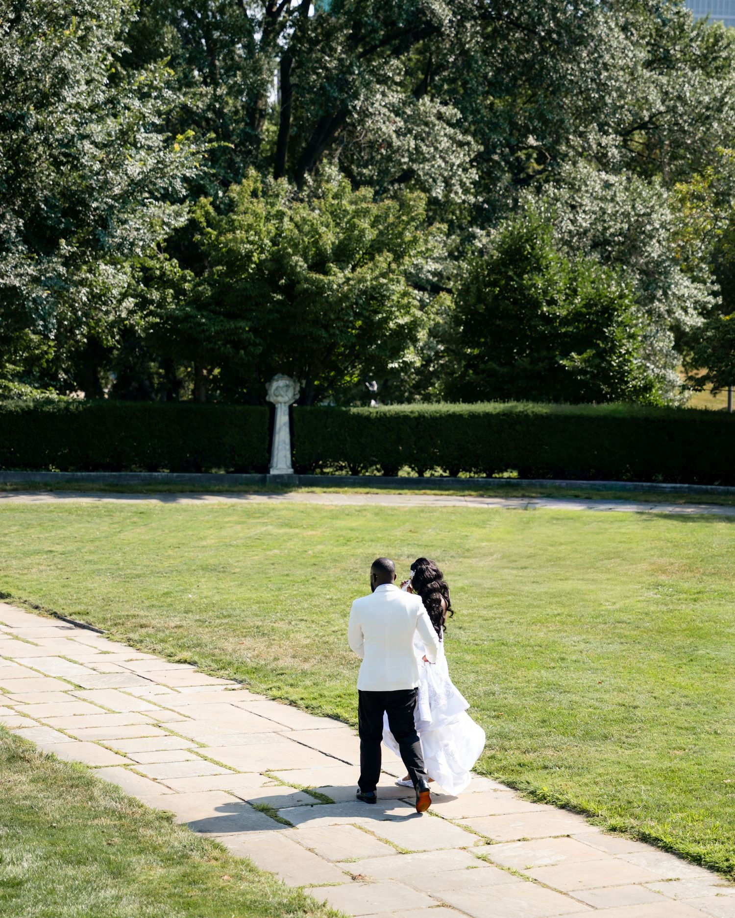 Bride and groom walk away along stone garden path surrounded by lush green hedges and trees.