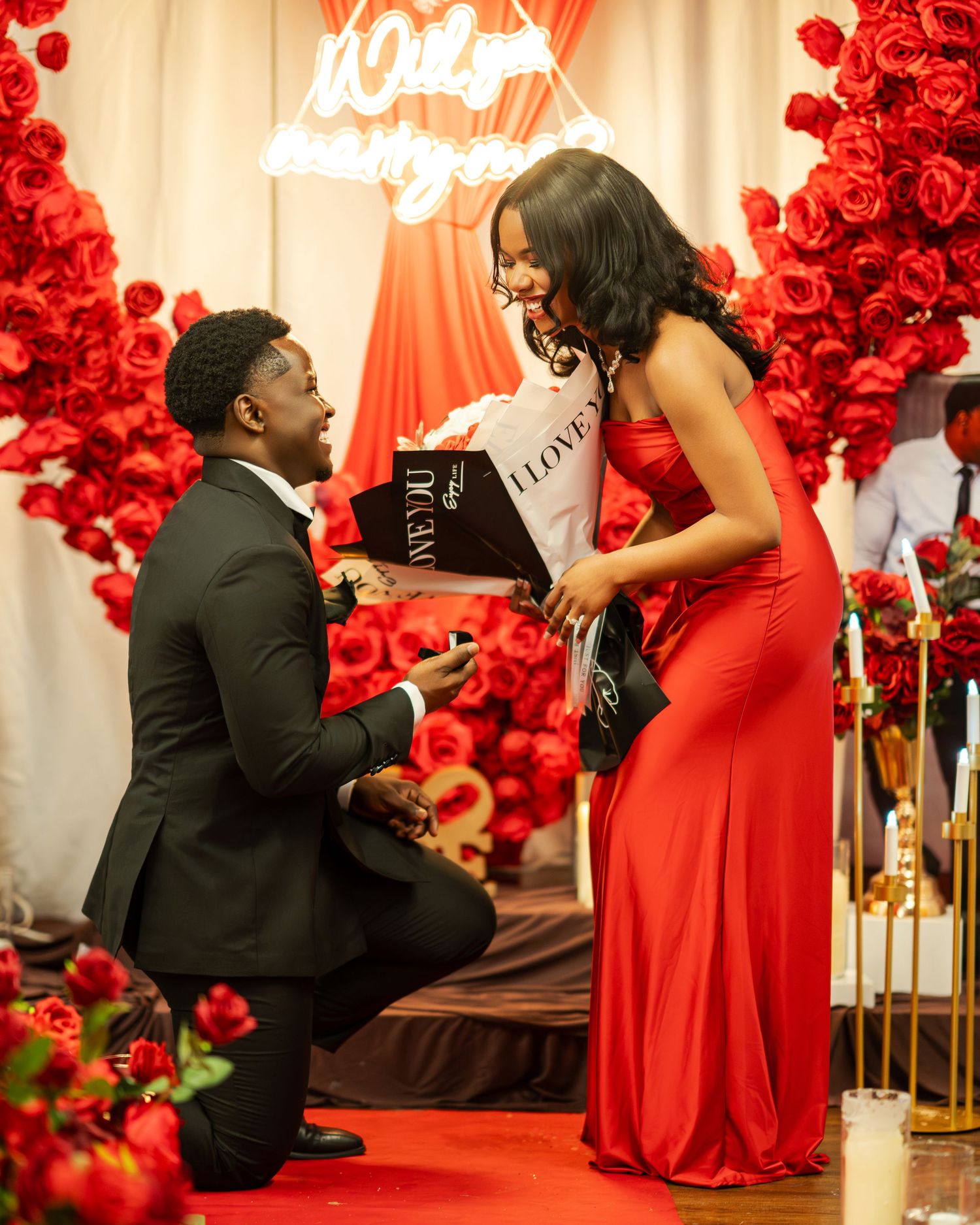 A romantic proposal scene with a man kneeling before a woman in a red gown holding an I Love You book.