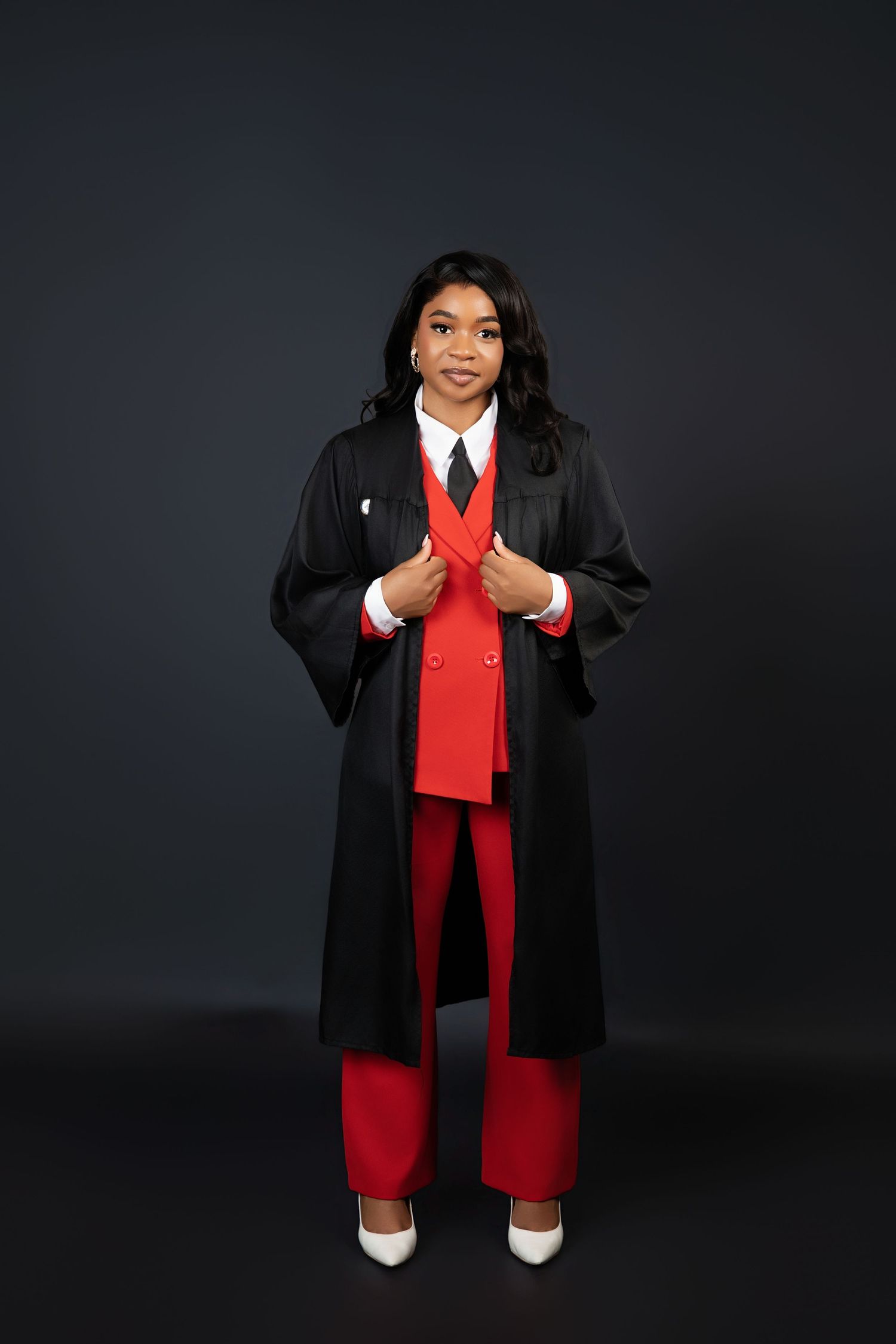 Graduate wearing black academic gown over a red suit with white heels against a dark background.