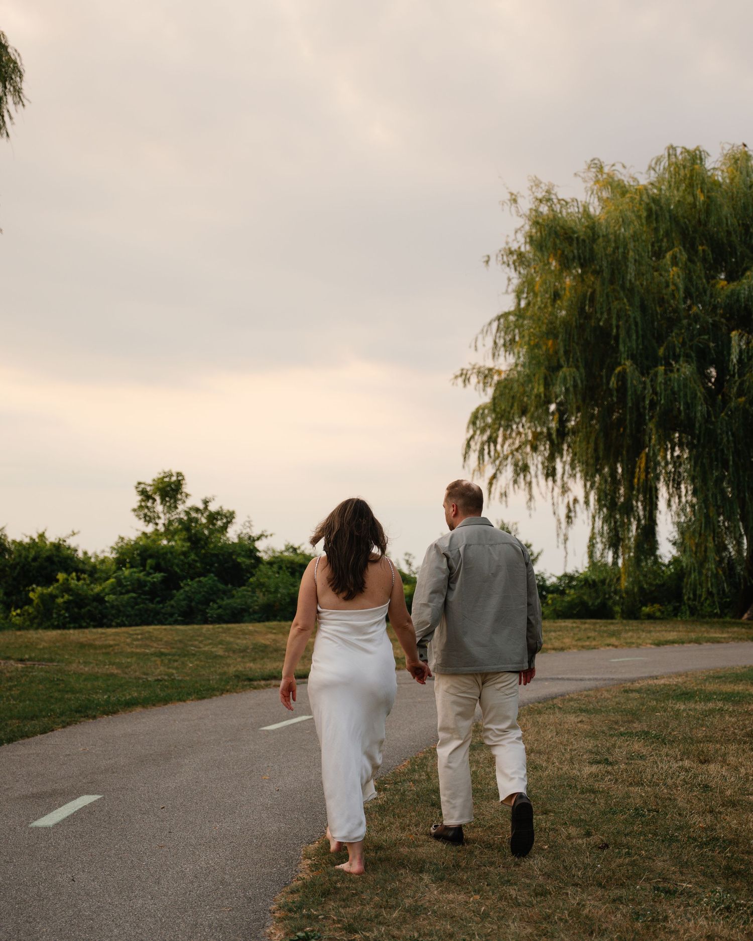 Couple holding hands walking down a road near a weeping willow tree at golden hour sunset.