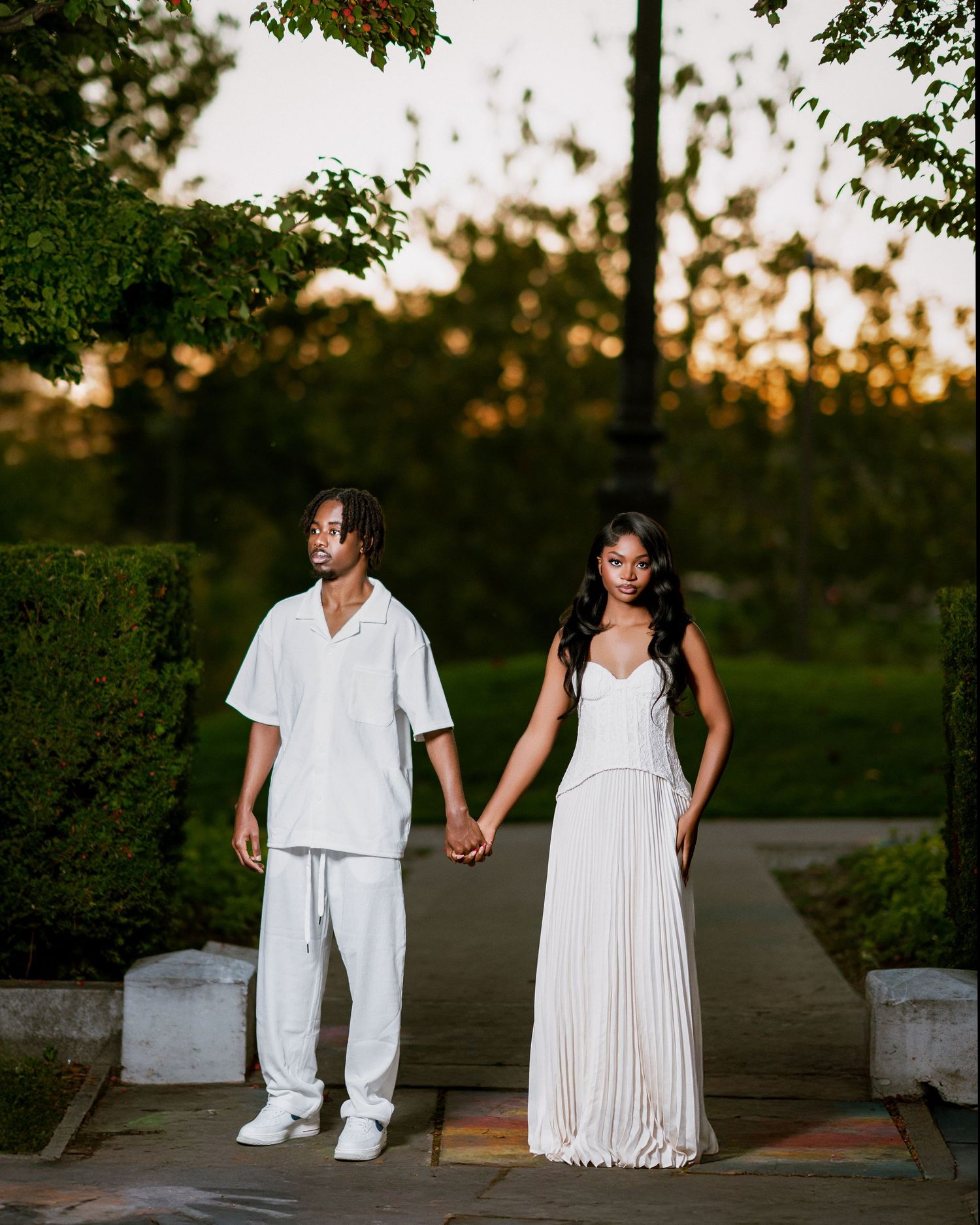 Couple dressed in white holding hands on a garden pathway during a golden sunset.
