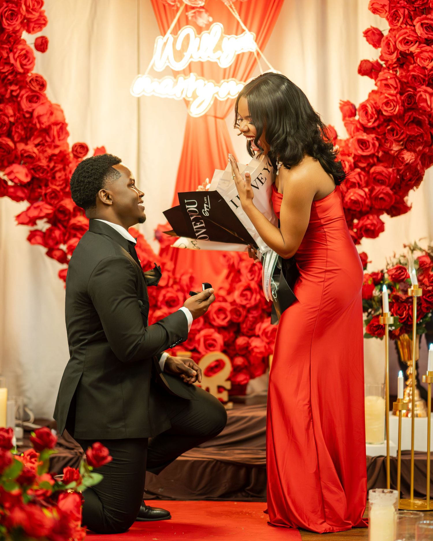 Man proposing to woman in red dress surrounded by red roses and romantic decor.