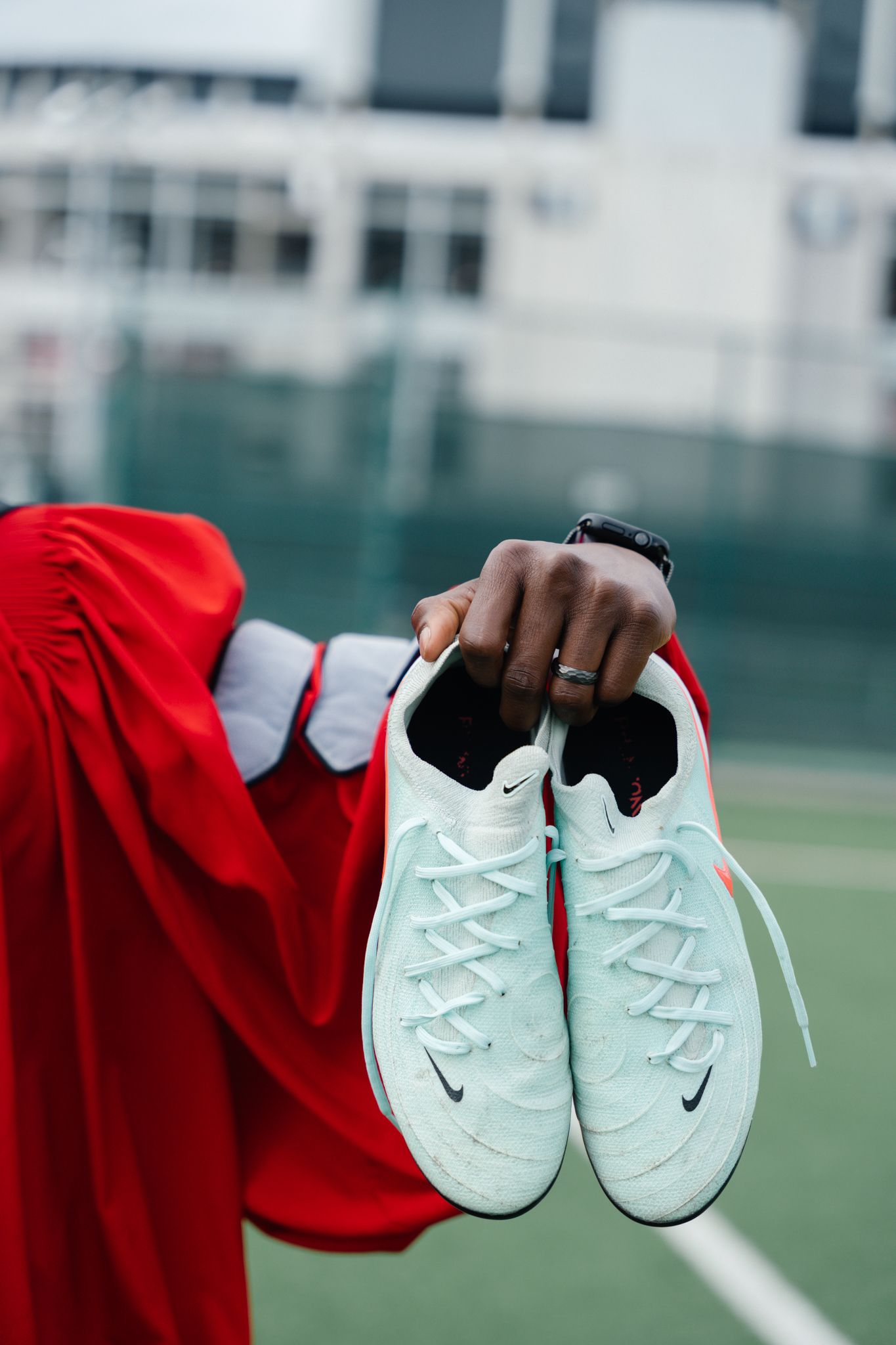 Hand holding white Nike sneakers wearing red graduation gown on athletic field.