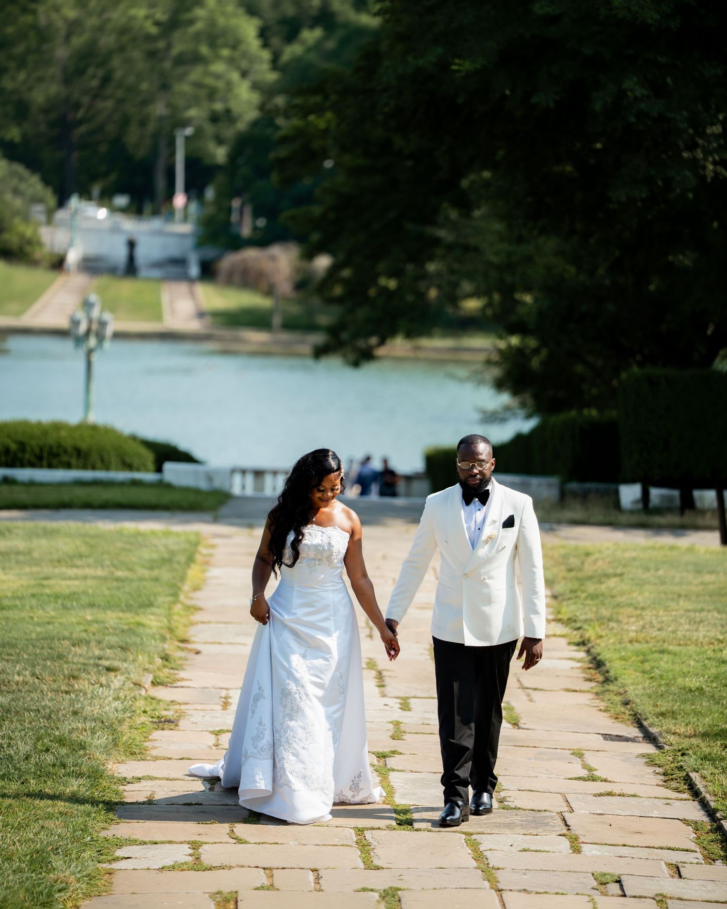 Bride and groom walk hand in hand along scenic tree-lined path near a reflecting pool.