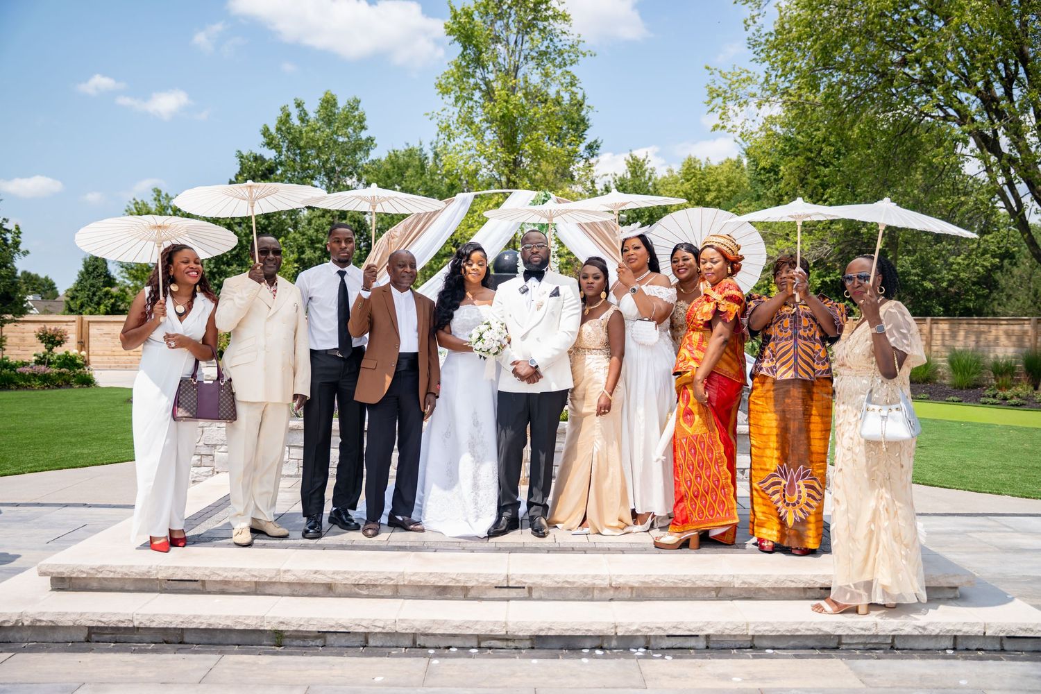 Wedding party group photo with bride and groom holding white parasols in sunny garden.