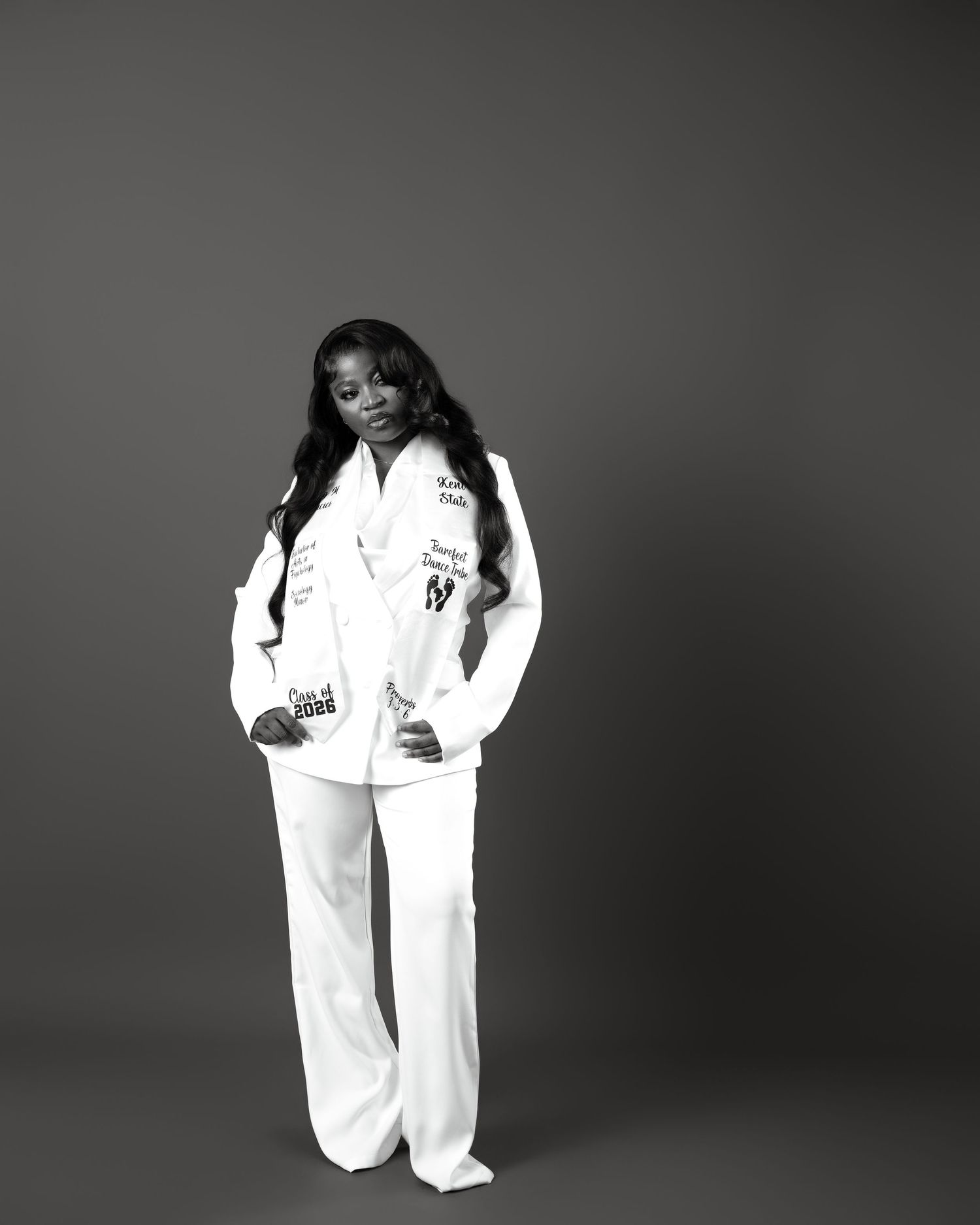 Black and white portrait of a woman in a white suit with patches standing against a gray background.