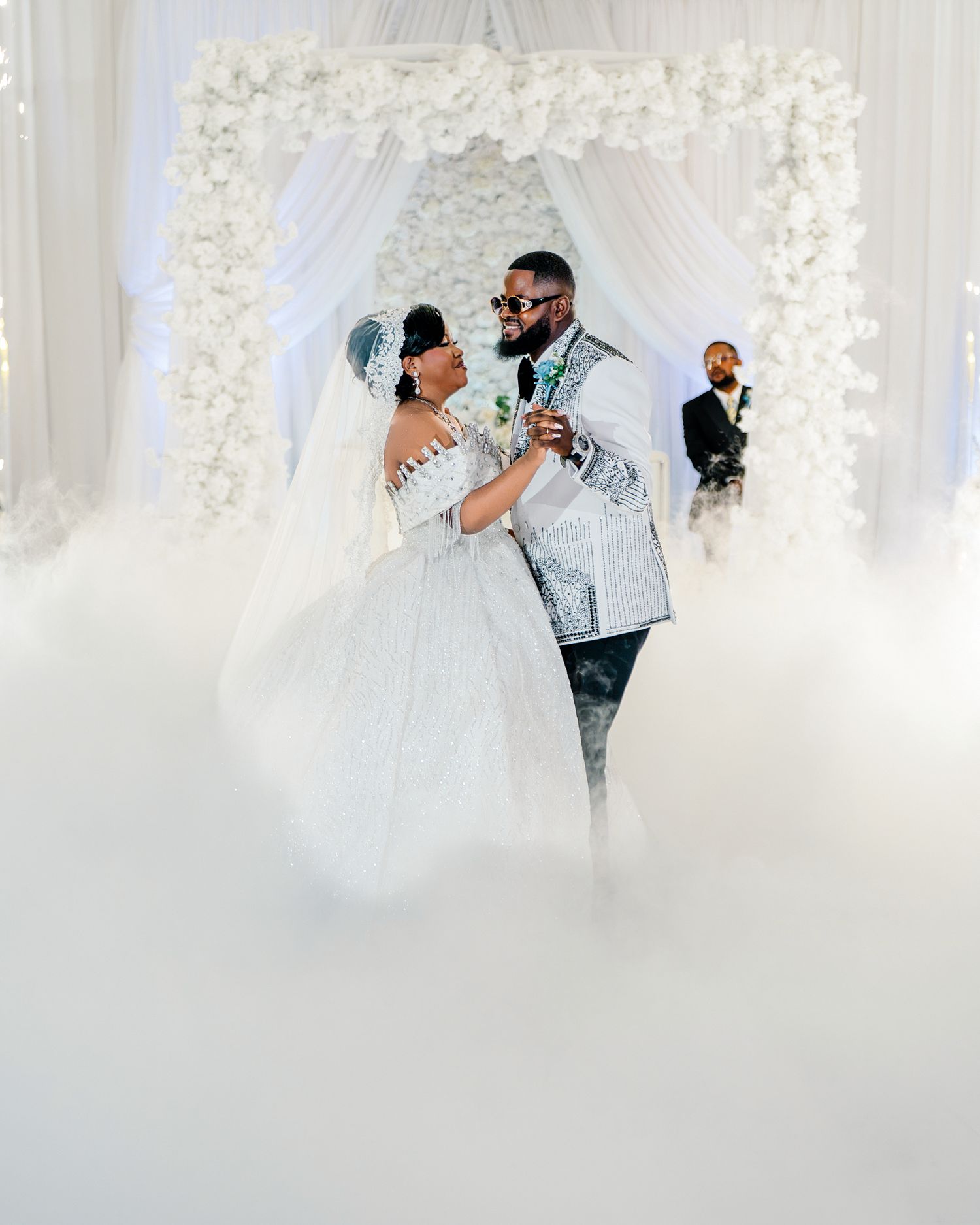 Bride and groom share first dance surrounded by dramatic fog under white floral wedding arch.