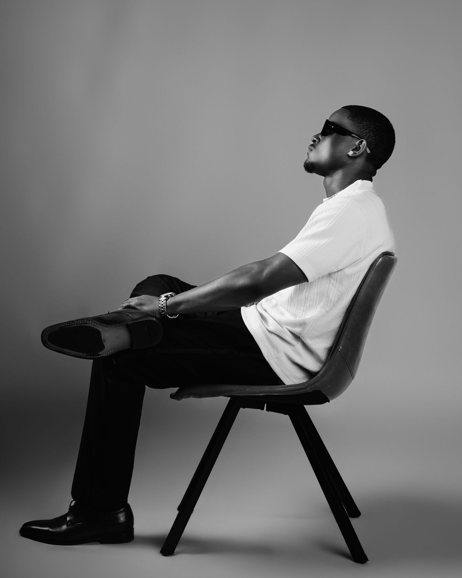 A Black man in a white shirt and black pants sits in a chair in a black and white studio portrait.