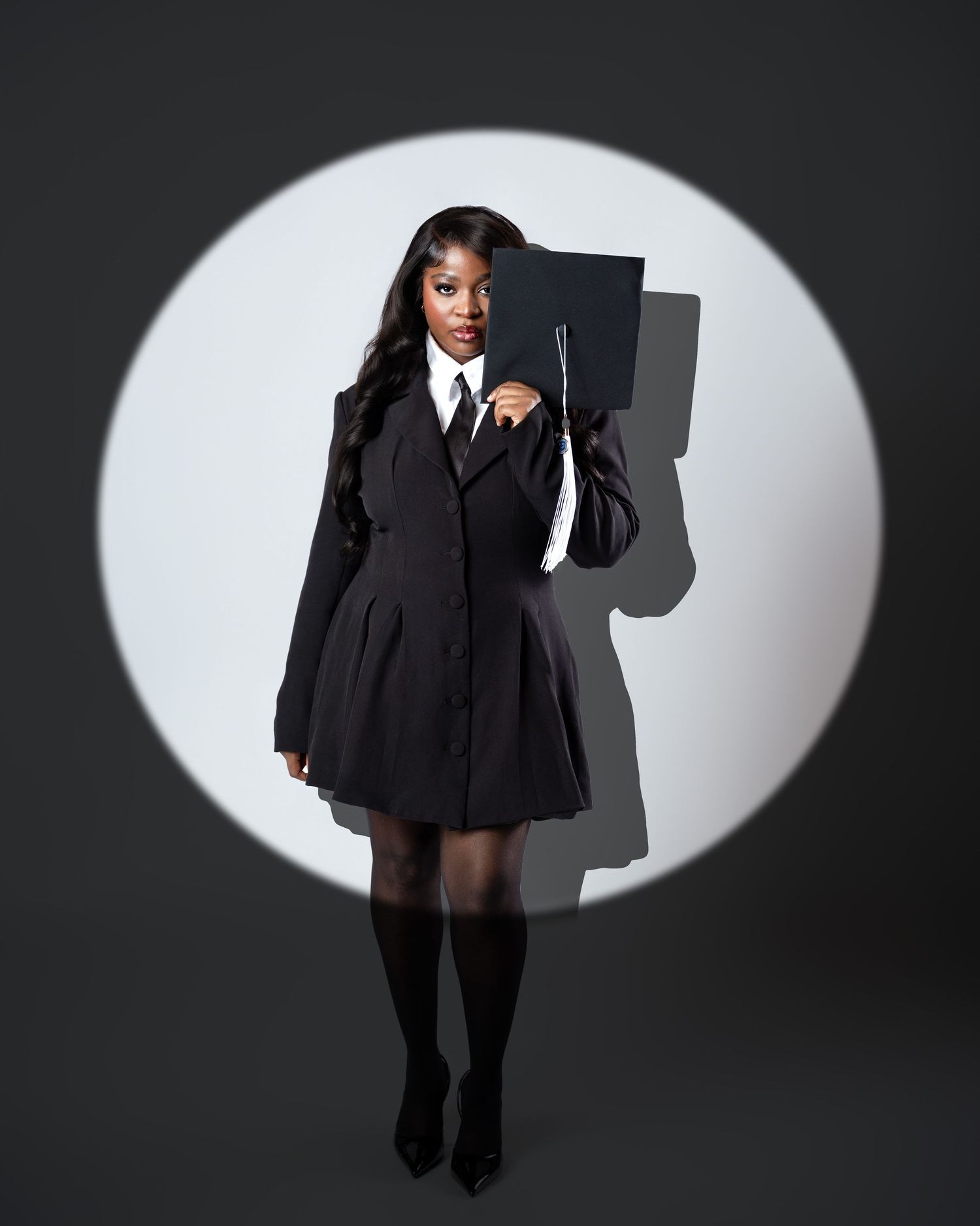A Black woman in a black blazer dress holds a graduation cap under a spotlight in a dramatic portrait.