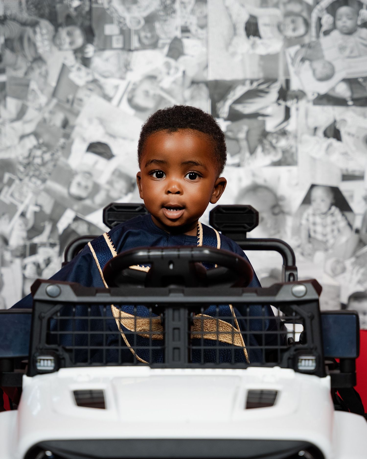 A cute toddler in a navy outfit sitting in a toy ride-on car against a black and white photo collage backdrop.