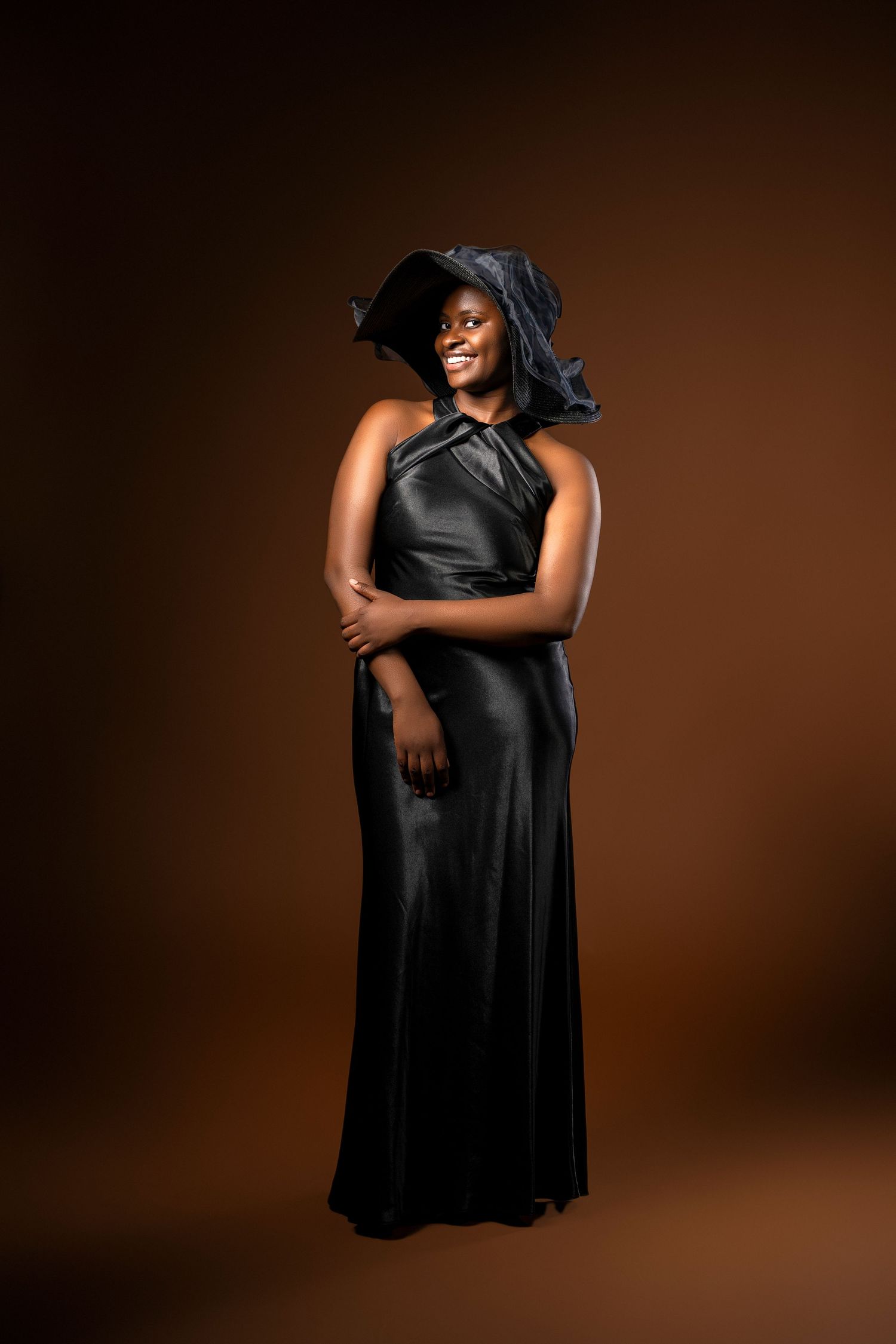 A smiling Black woman poses in a black satin halter gown and wide-brim hat against a brown background.