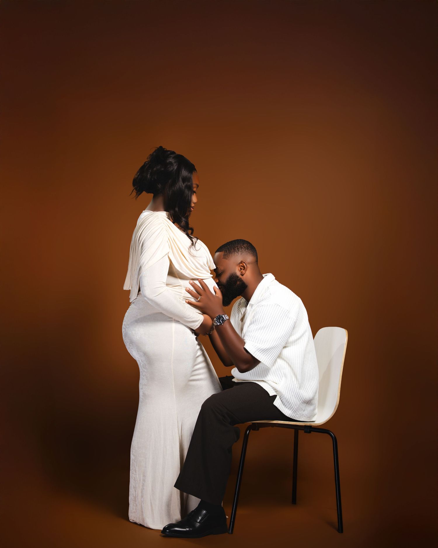 Maternity photo of a couple in white outfits, man kisses pregnant woman's belly on brown background.