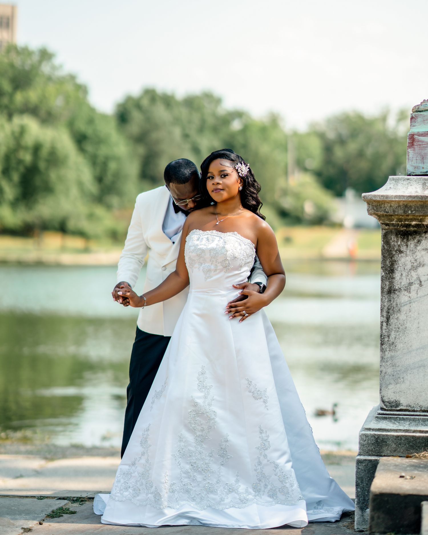 Wedding couple poses by lakeside, bride in white gown and groom in white tuxedo jacket.