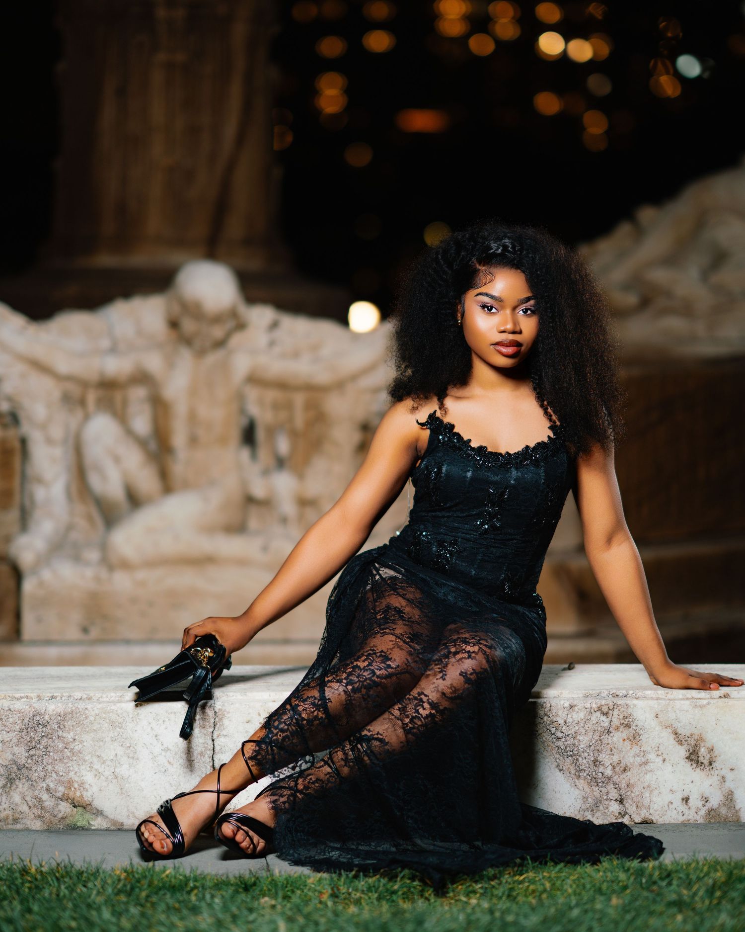 Elegant young woman in black lace gown seated on stone steps at night with ornate sculpture backdrop.