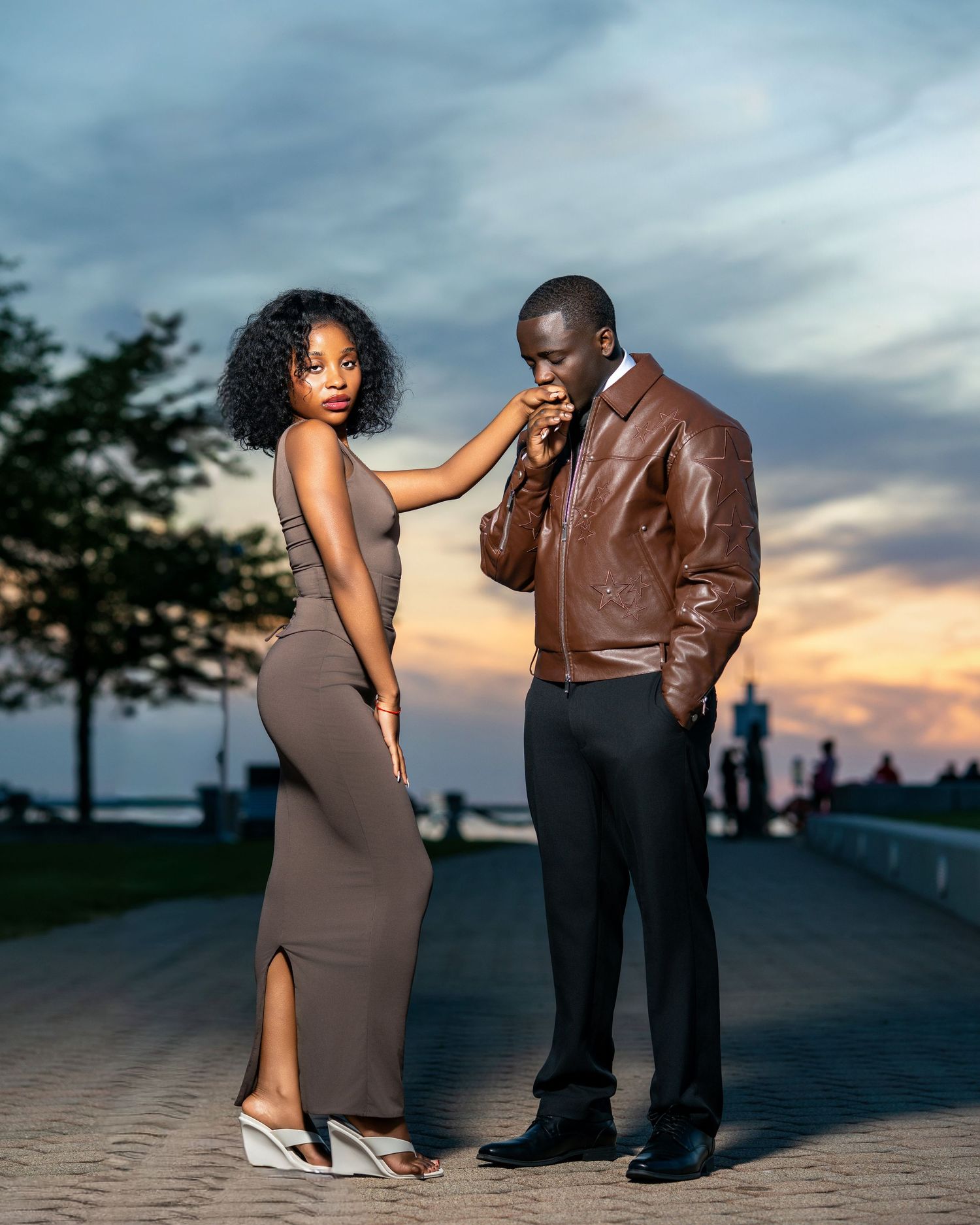 Couple at sunset waterfront, man kissing woman's hand, both wearing brown and grey outfits.