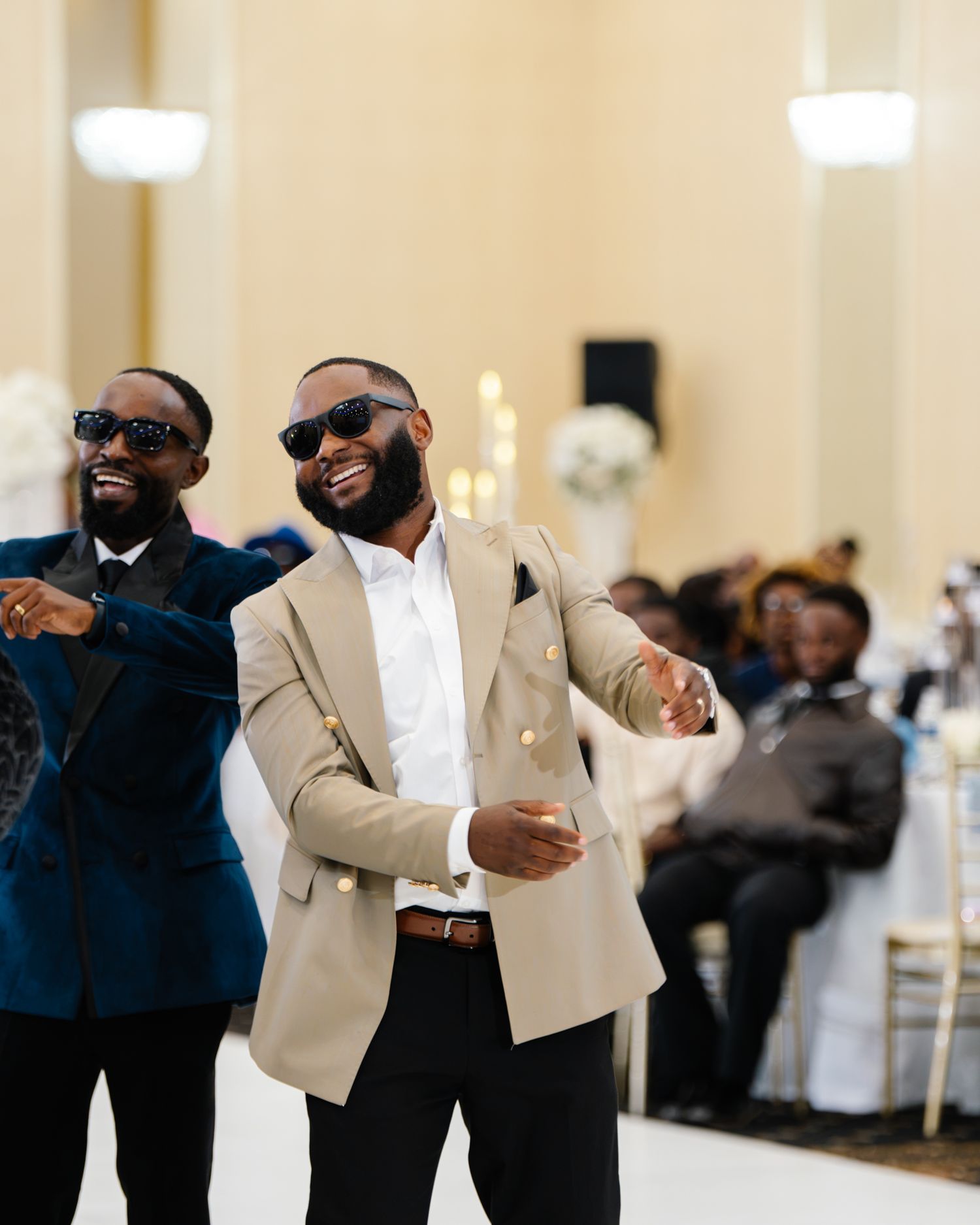 Two well-dressed men in suits dance and celebrate at an elegant wedding reception.