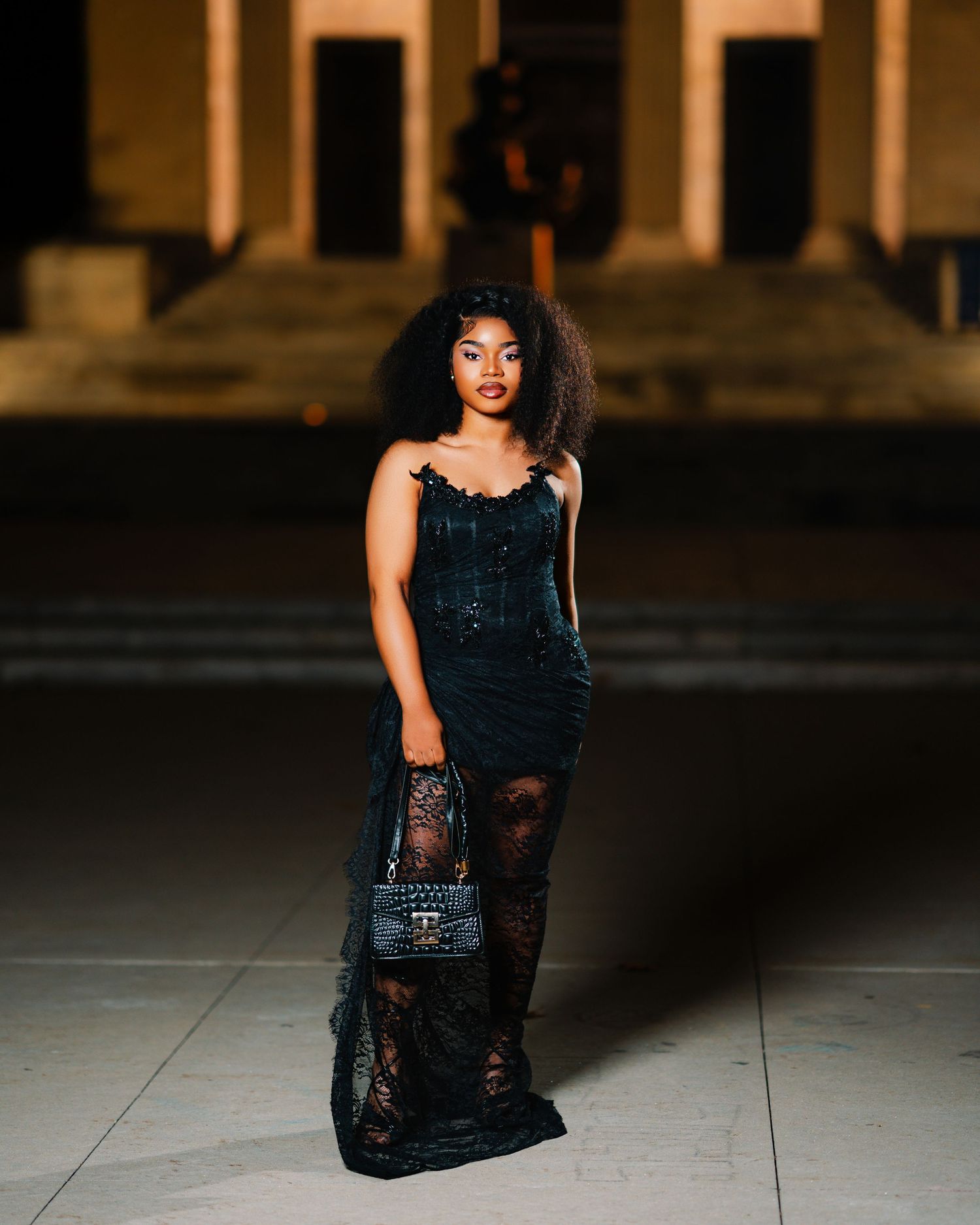 Fashionable young woman in black lace dress holding designer handbag posing on plaza steps at night.
