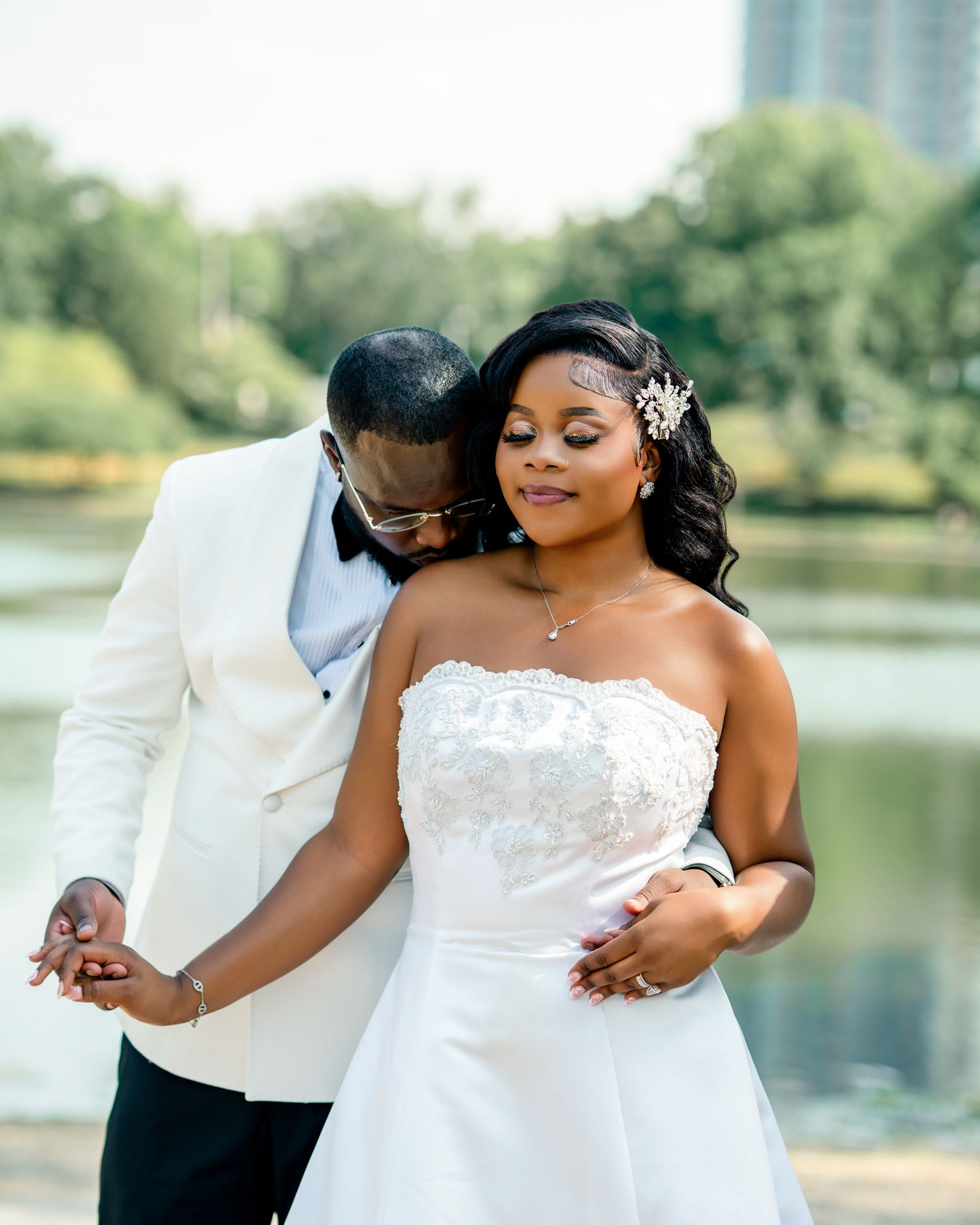 A romantic bride and groom embrace near a serene lake, with the groom nuzzling the bride in her white strapless gown.