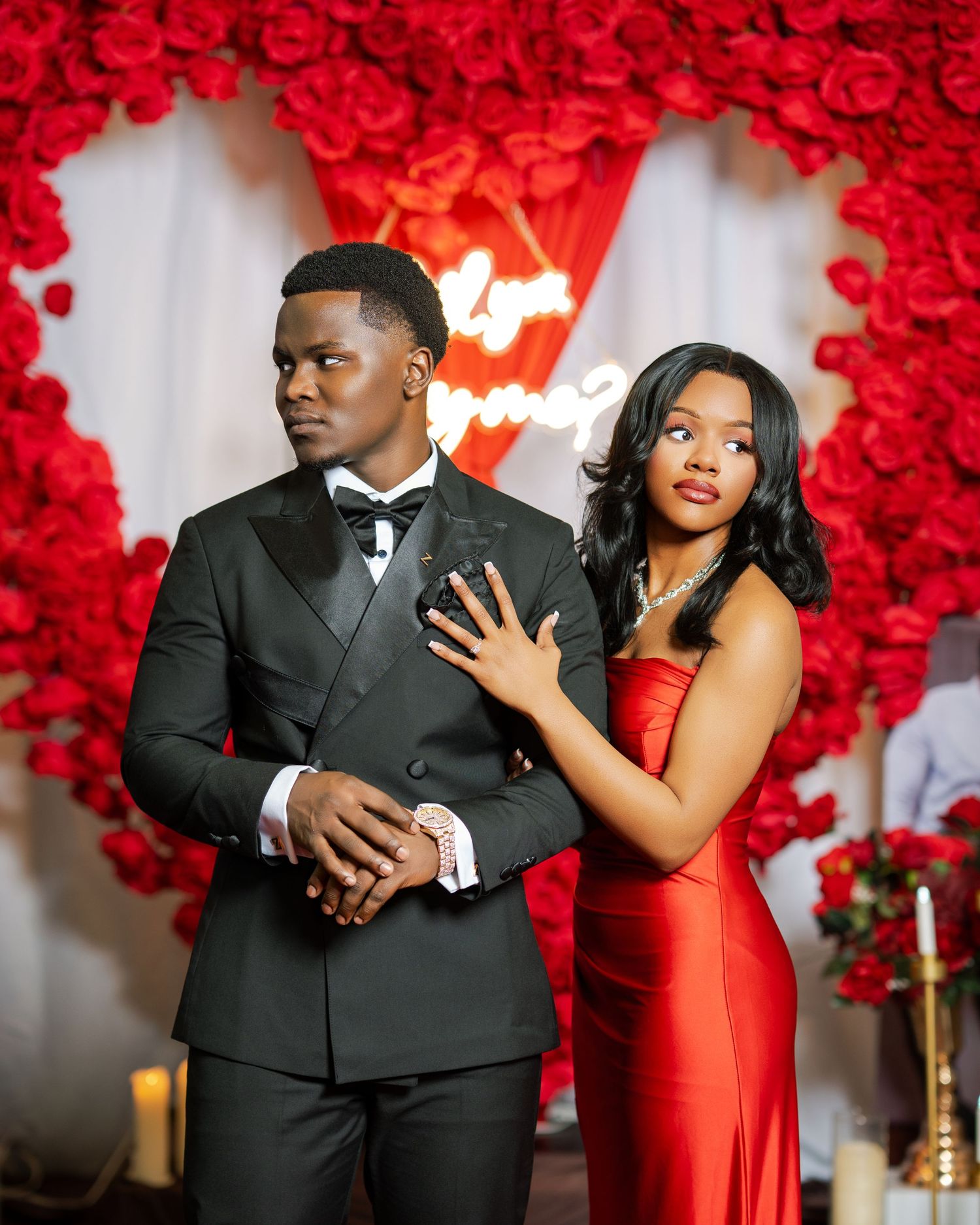 An engaged couple poses before a heart-shaped red rose wall with a glowing Will You Marry Me neon sign.