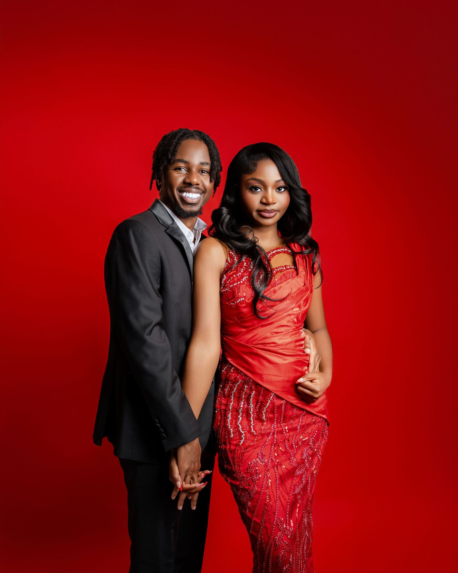 A stylish Black couple poses against a red background, wearing elegant red dress and black suit.