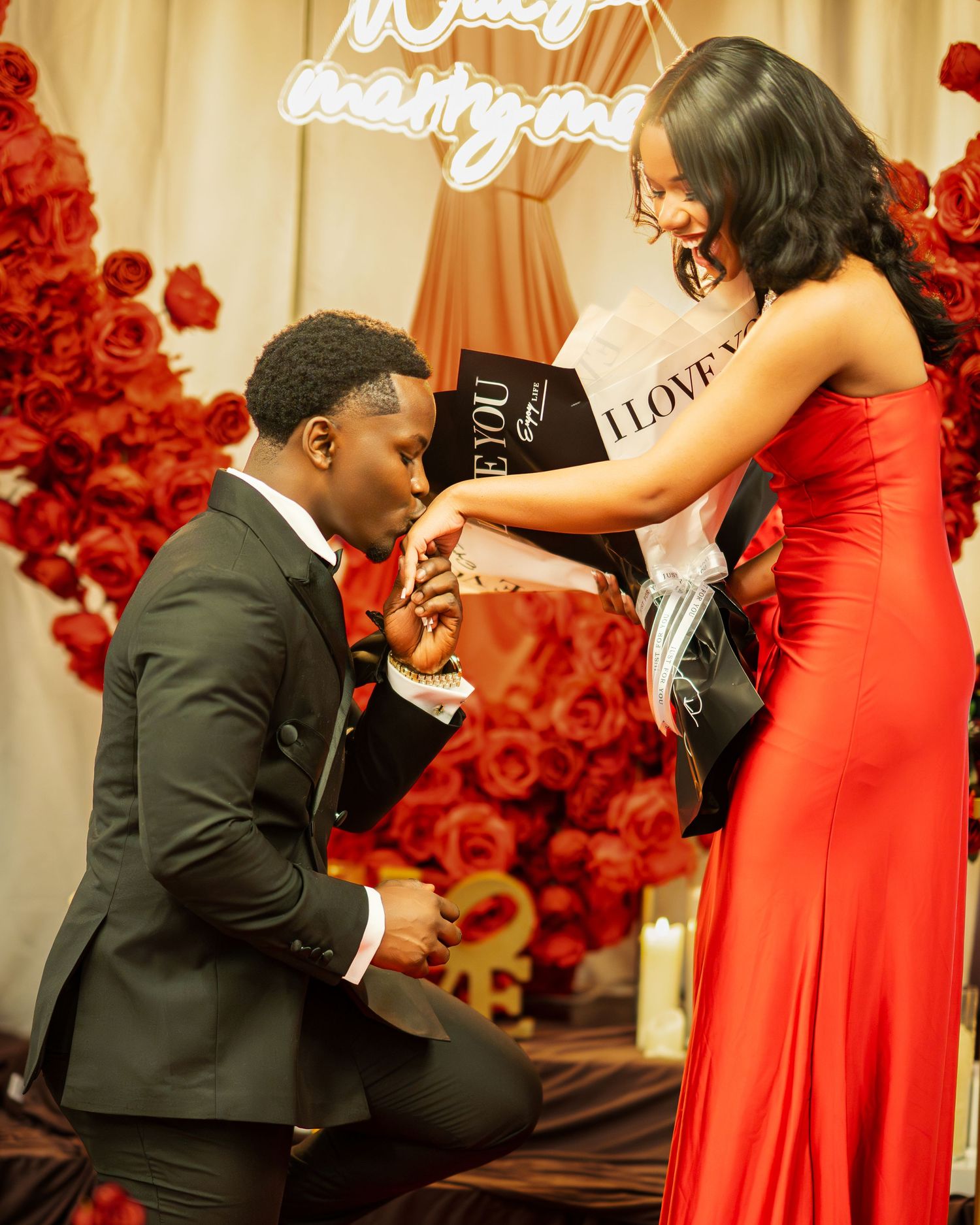 A man on one knee places a ring on a woman's finger during a stunning red rose-decorated marriage proposal.