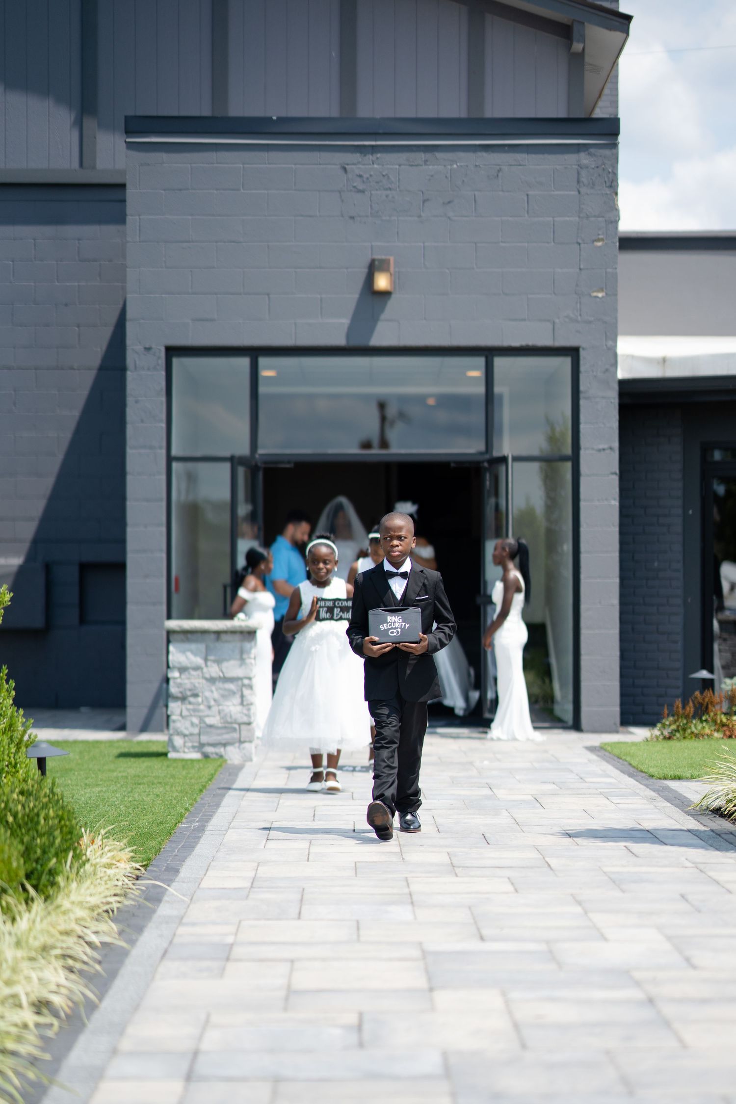 Young ring bearer and flower girl leading wedding processional down stone pathway from modern venue.