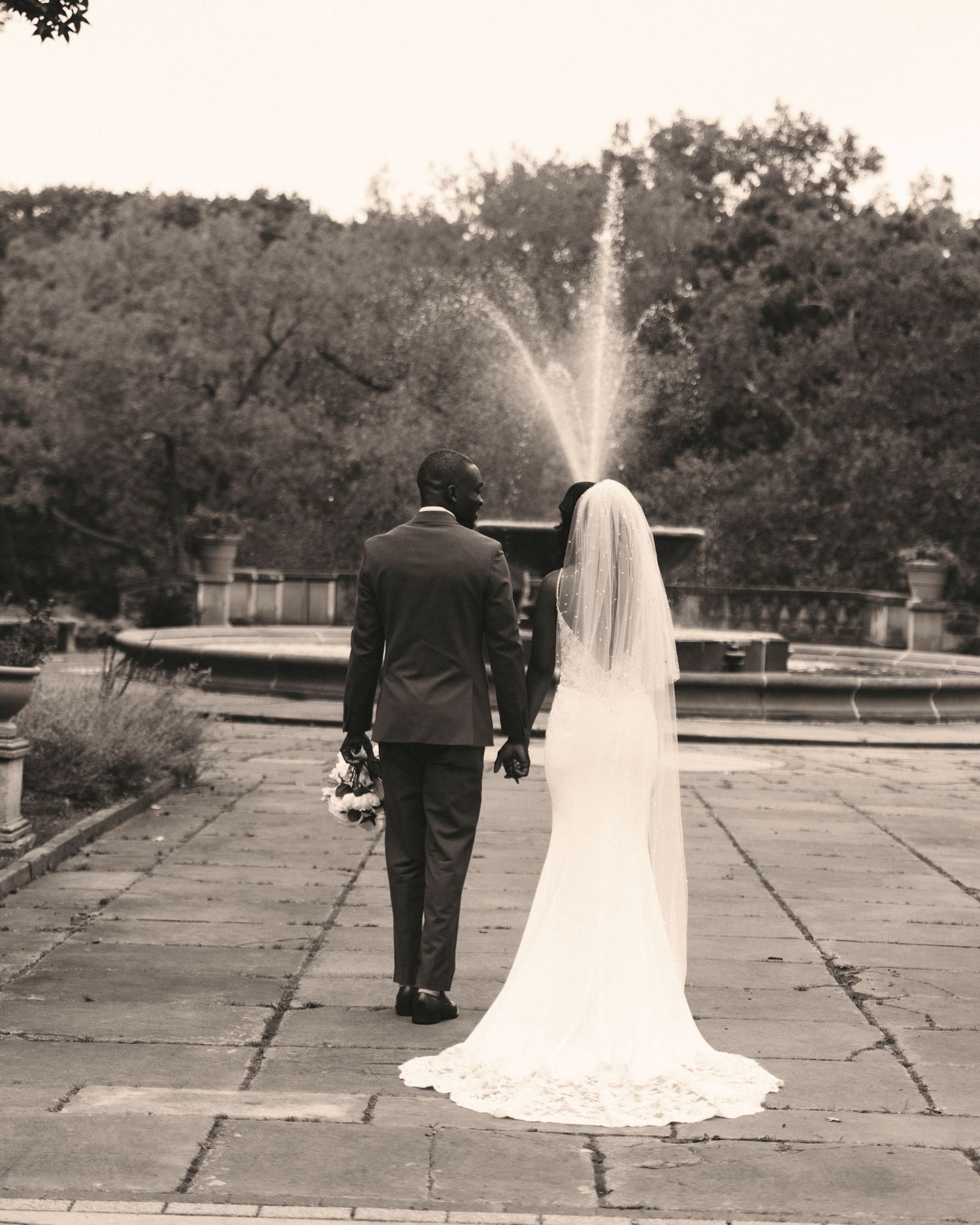 A bride and groom walk hand in hand toward a fountain in a black and white wedding photo.