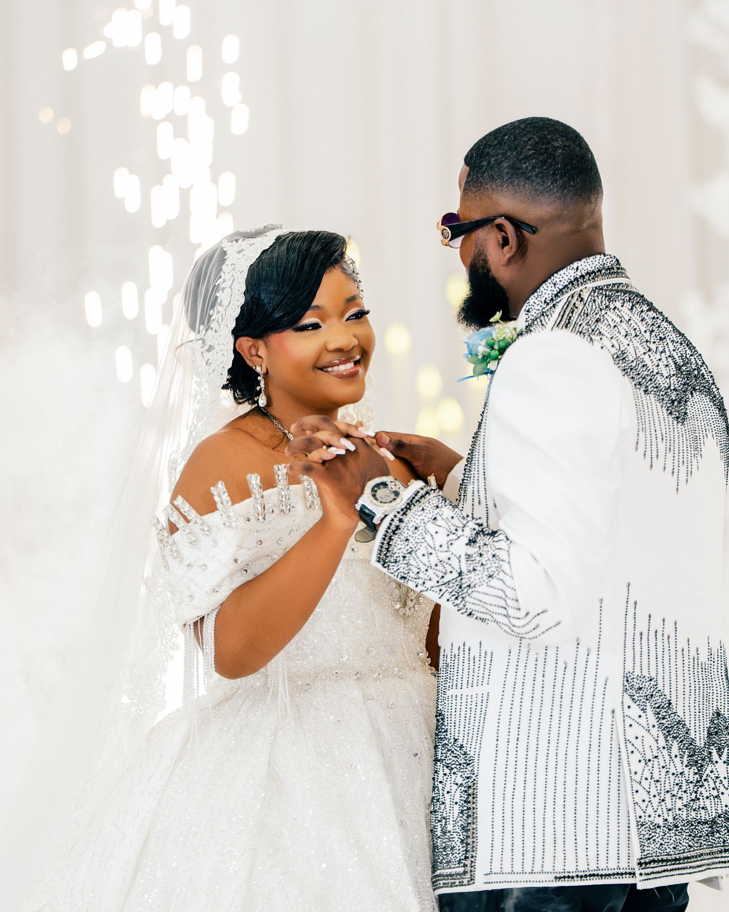 Bride in beaded gown and groom in embellished jacket sharing intimate moment at wedding.