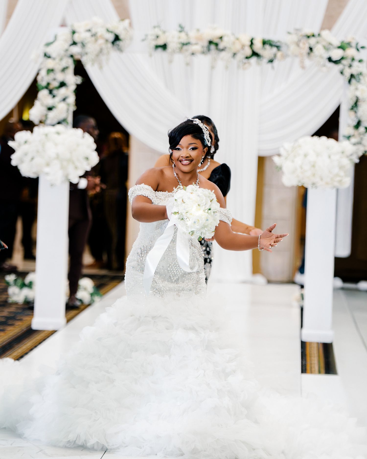 Joyful bride in a white mermaid wedding gown with feather hem walks down the aisle holding a bouquet.
