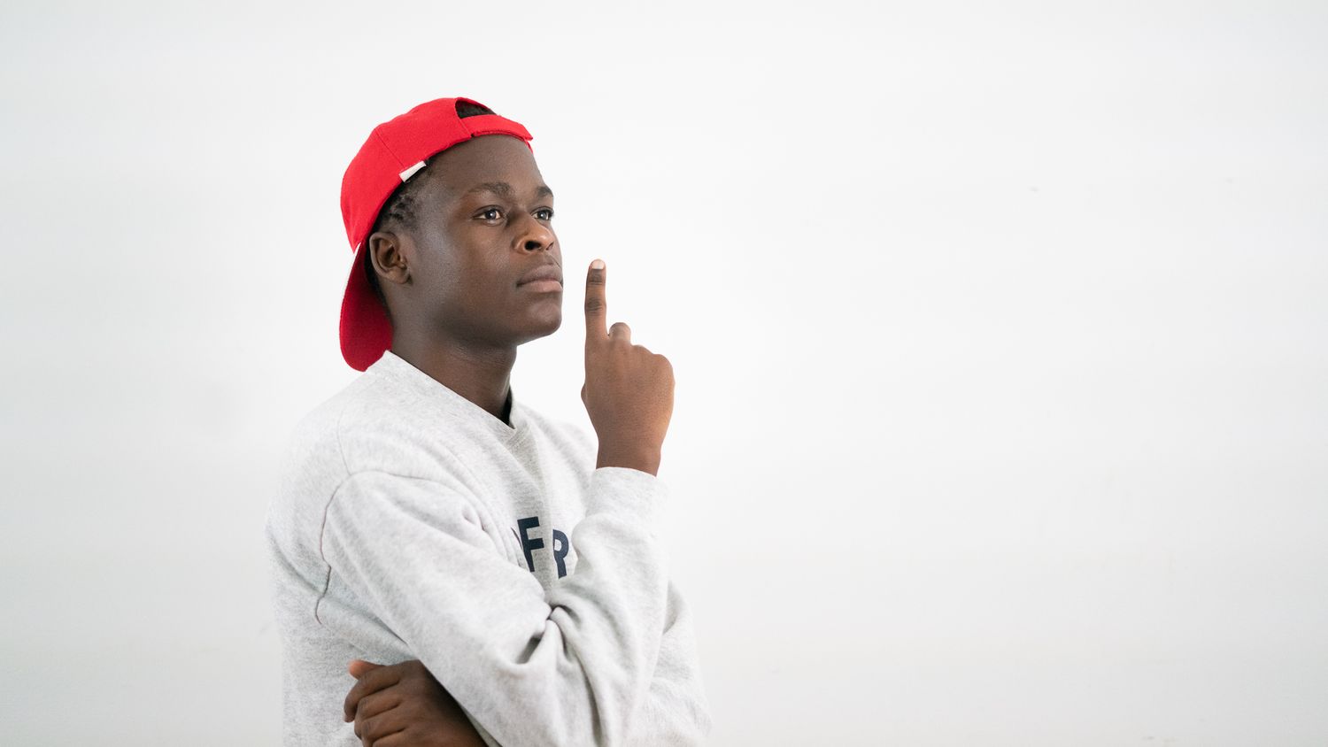 Young man in a red cap and white hoodie pointing upward against a white background.